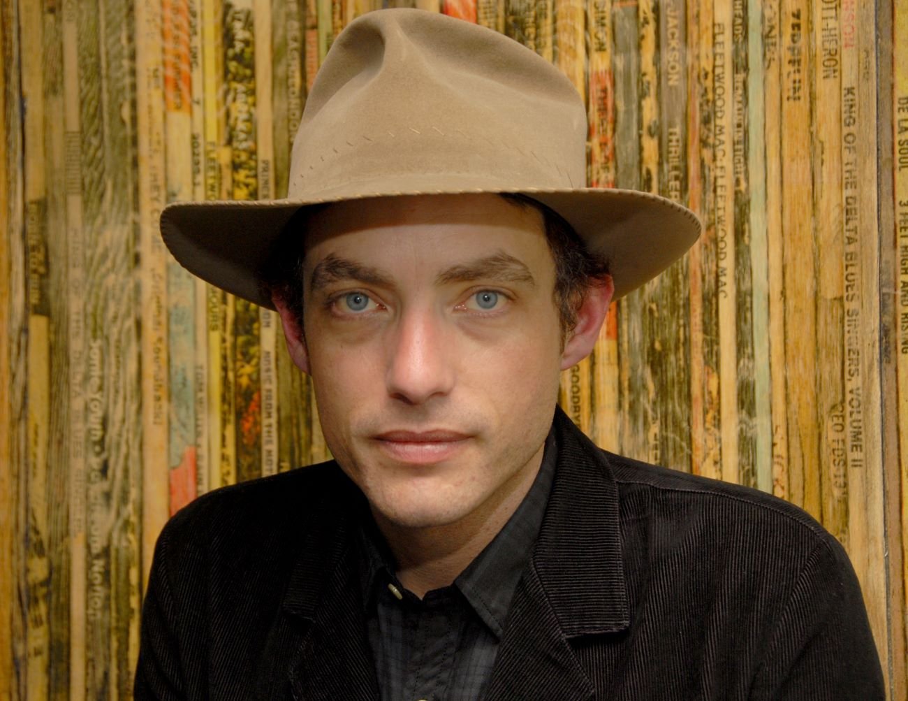 Jakob Dylan wears a hat and stands in front of a wall.