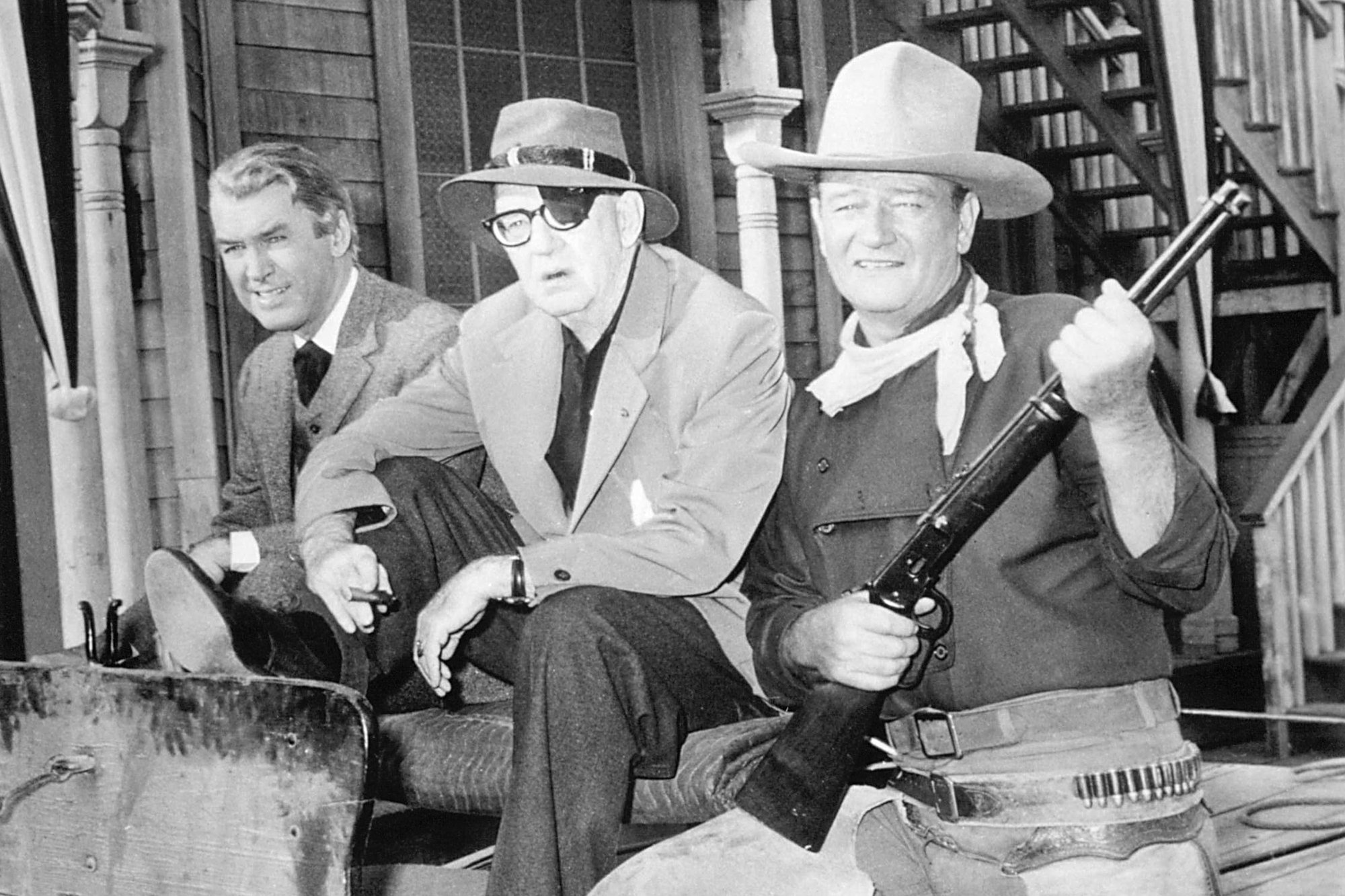 James Stewart, John Ford, and John Wayne sitting on a horse carriage in a black and white photo