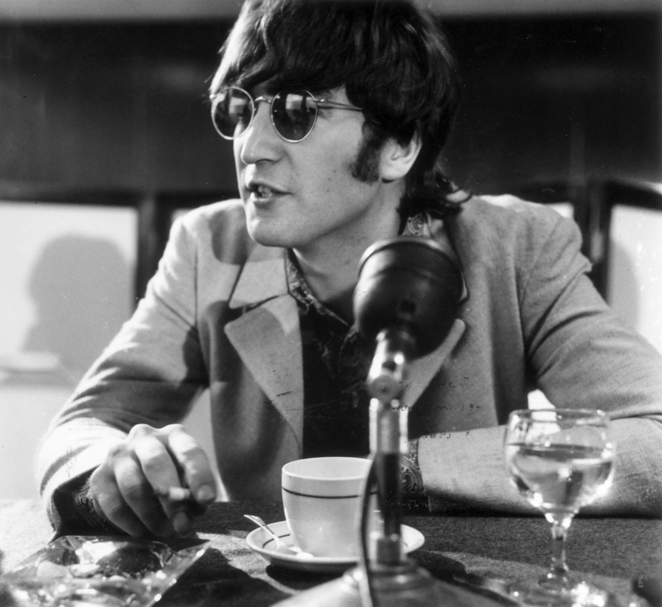 John Lennon wears sunglasses and sits in front of a microphone.