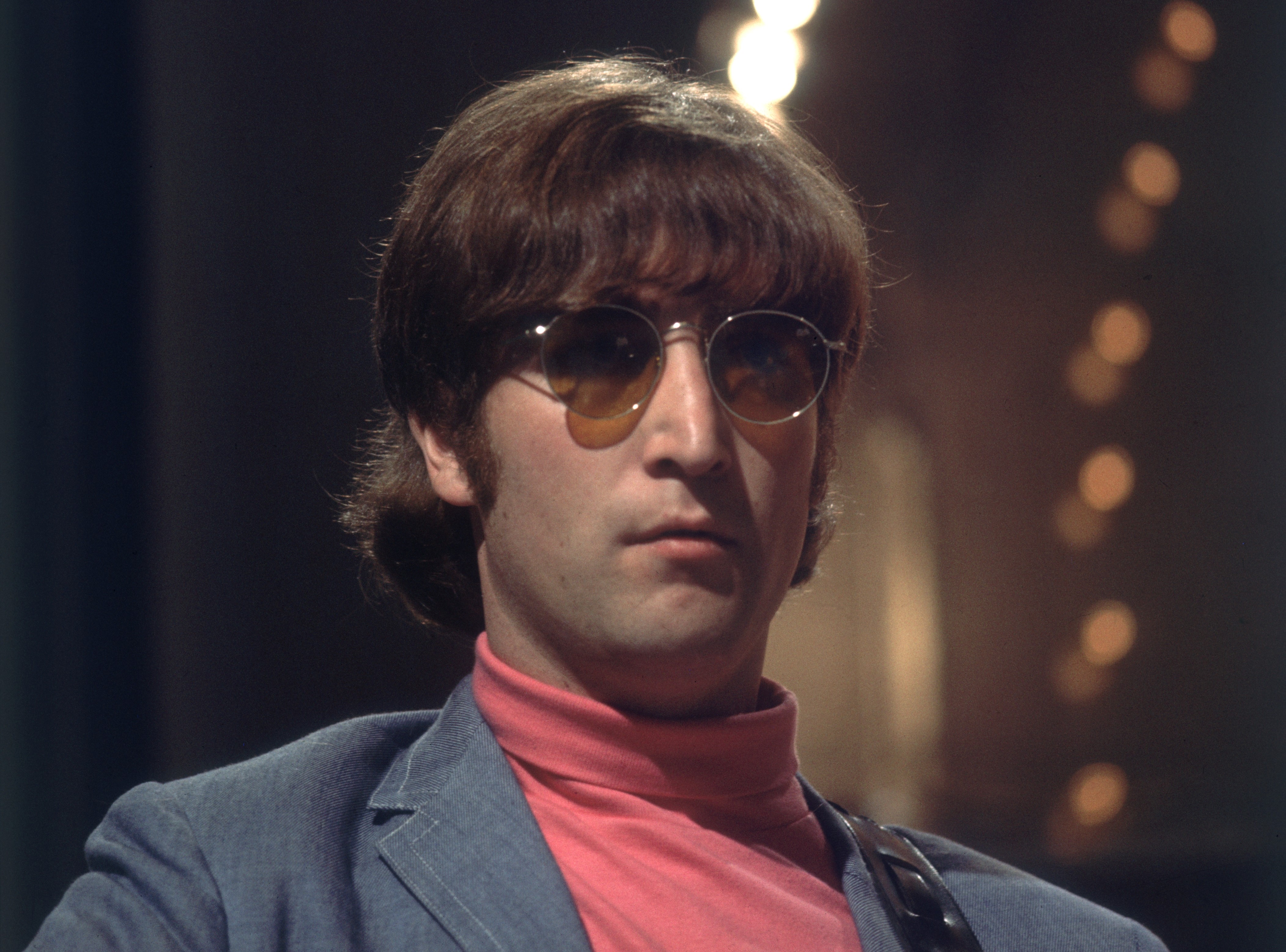 John Lennon wears a red turtleneck, jacket, and sunglasses.