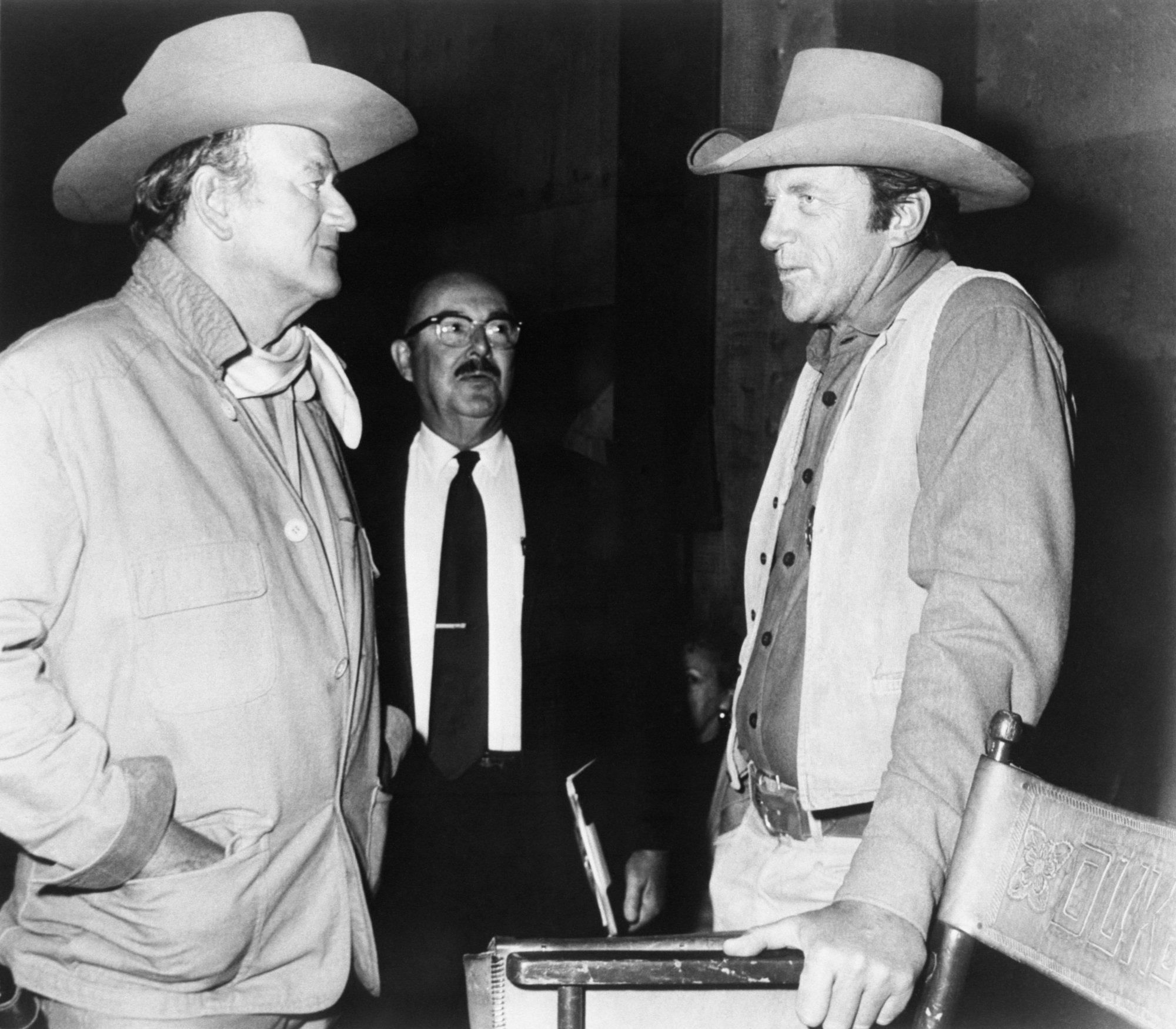 John Wayne and 'Gunsmoke' actor Jim Arness. They are facing one another wearing Western cowboy costumes. Wayne has his hands in his pockets and Arness rests his hand on a chair.