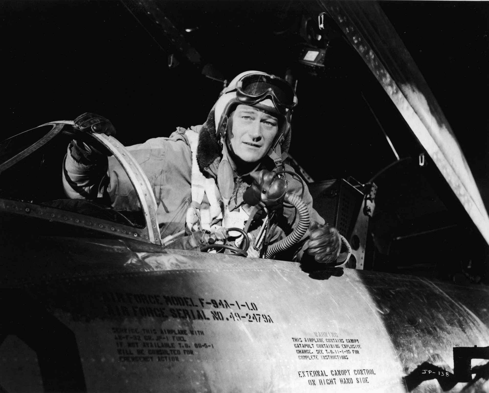 John Wayne as Col. Jim Shannon in 'Jet Pilot,' one of his worst movies. He's in a pilot uniform, sitting in the cockpit.