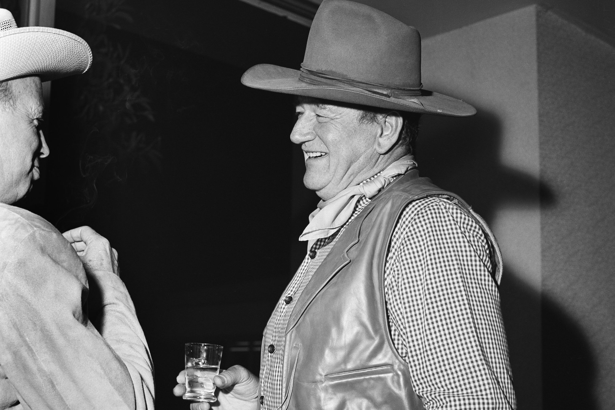 John Wayne holding a cocktail. He's smiling while wearing a cowboy hat and costume, talking to another party guest.