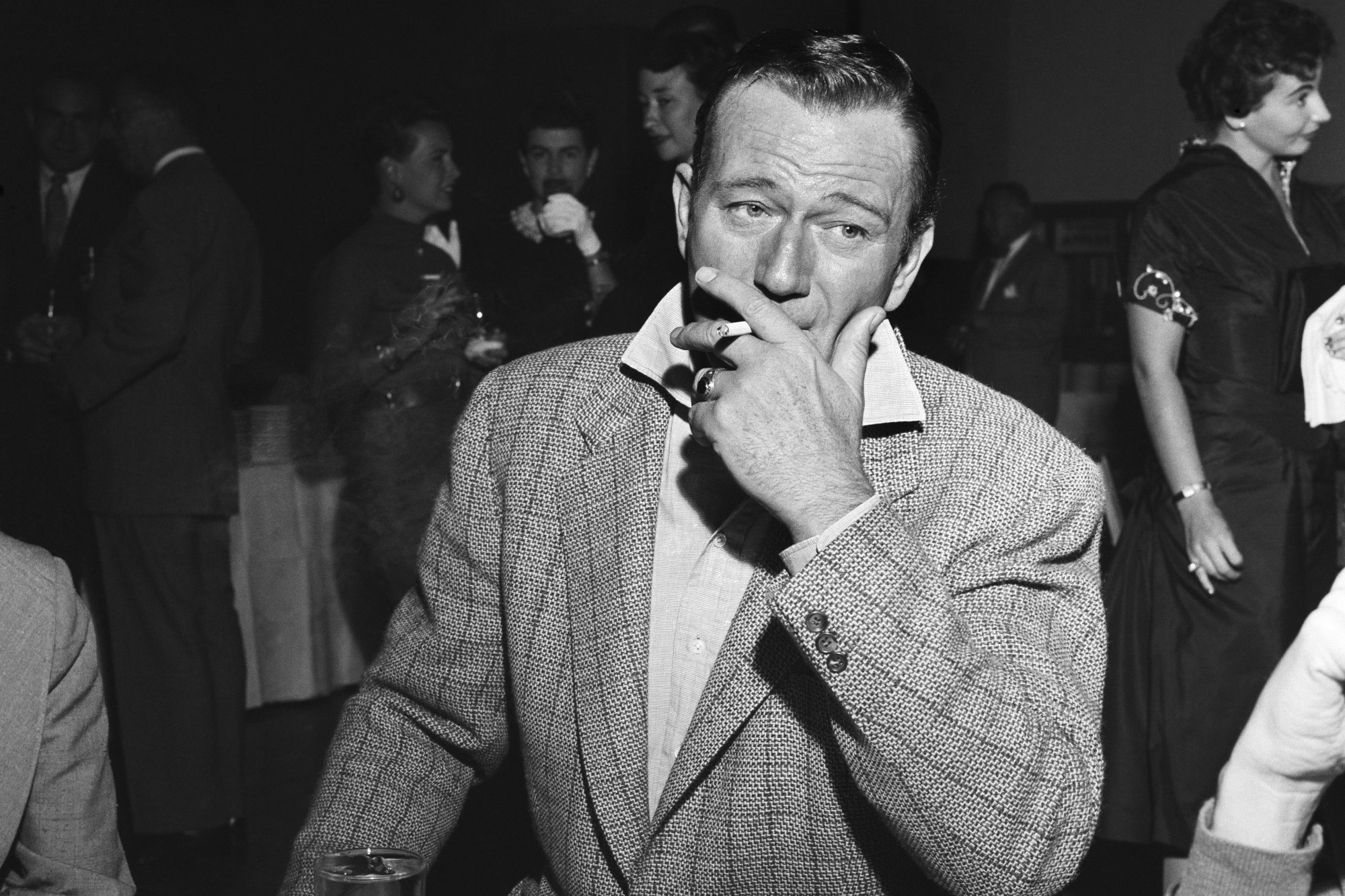 John Wayne, who co-created a club. He's wearing a suit and smoking a cigarette with a bunch of people at the party in the background.