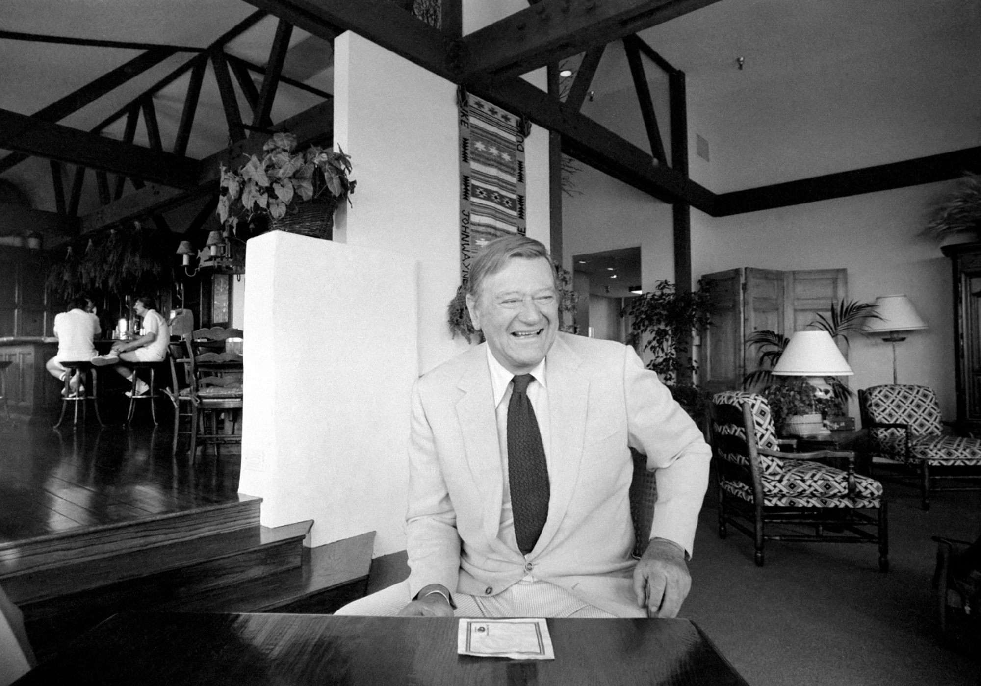 John Wayne, who has a coffee recipe made after him. He's smiling, wearing a suit and tie, seated at a table in his home.