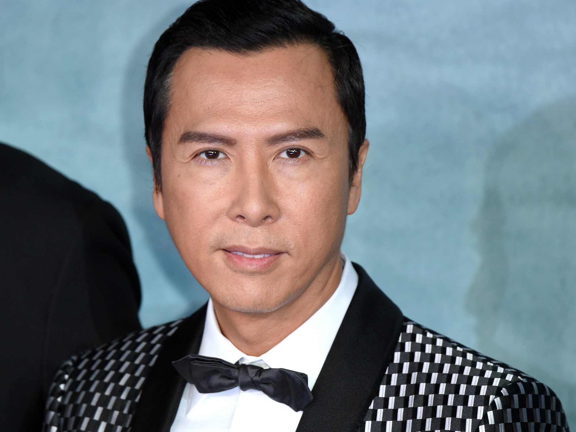 'John Wick 4' actor Donnie Yen wearing a black and white suit in front of a blue background