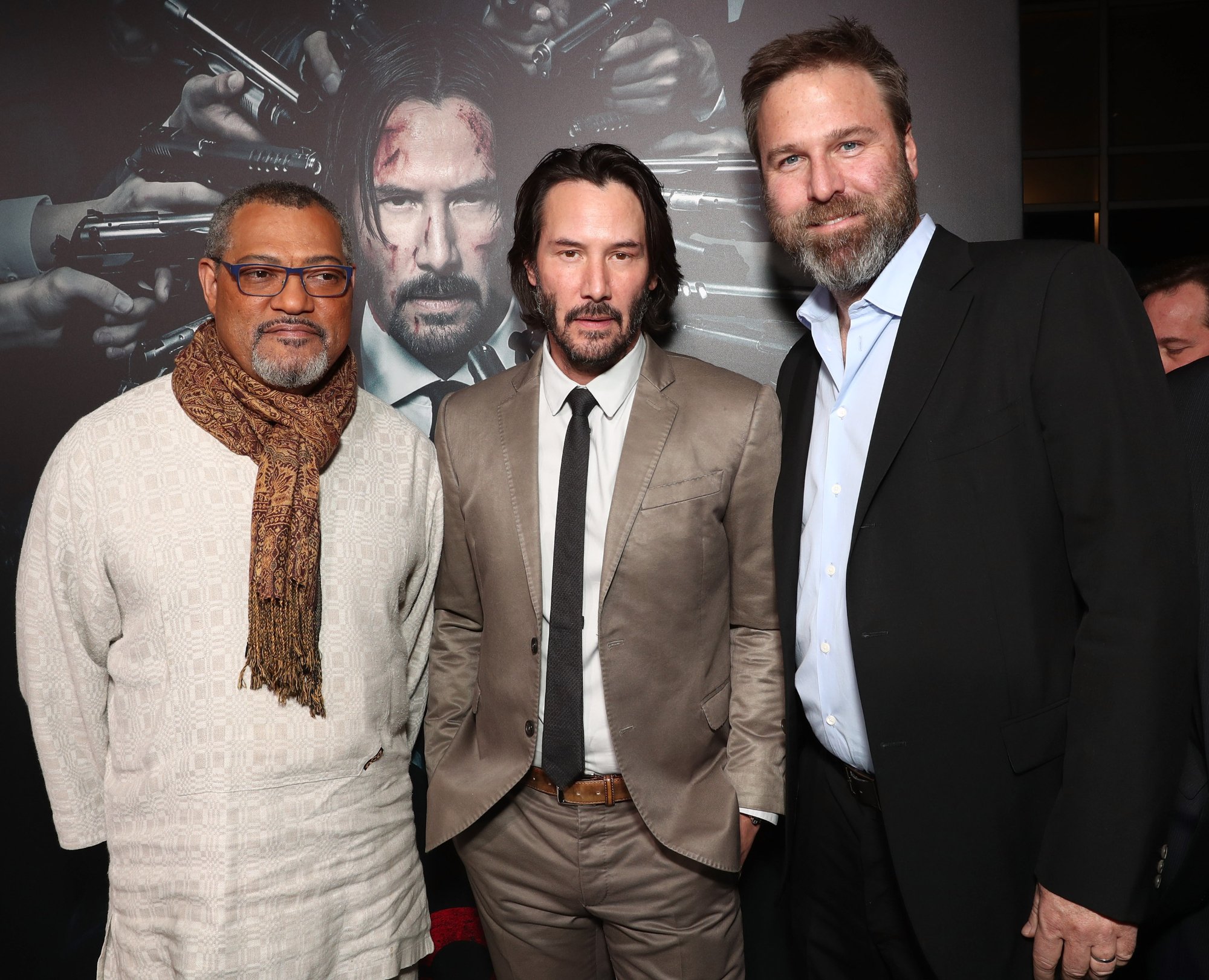 'John Wick 4' movie producer Basil Iwanyk and actors Laurence Fishburne and Keanu Reeves wearing suits in front of 'John Wick 2' poster