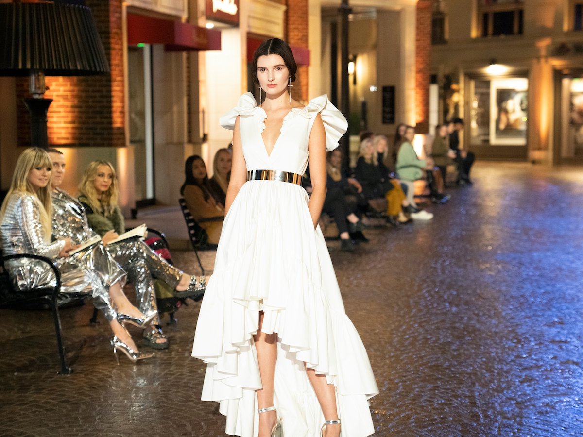A model wearing a white, high-low dress with a gold belt in 'Making the Cut' Season 3