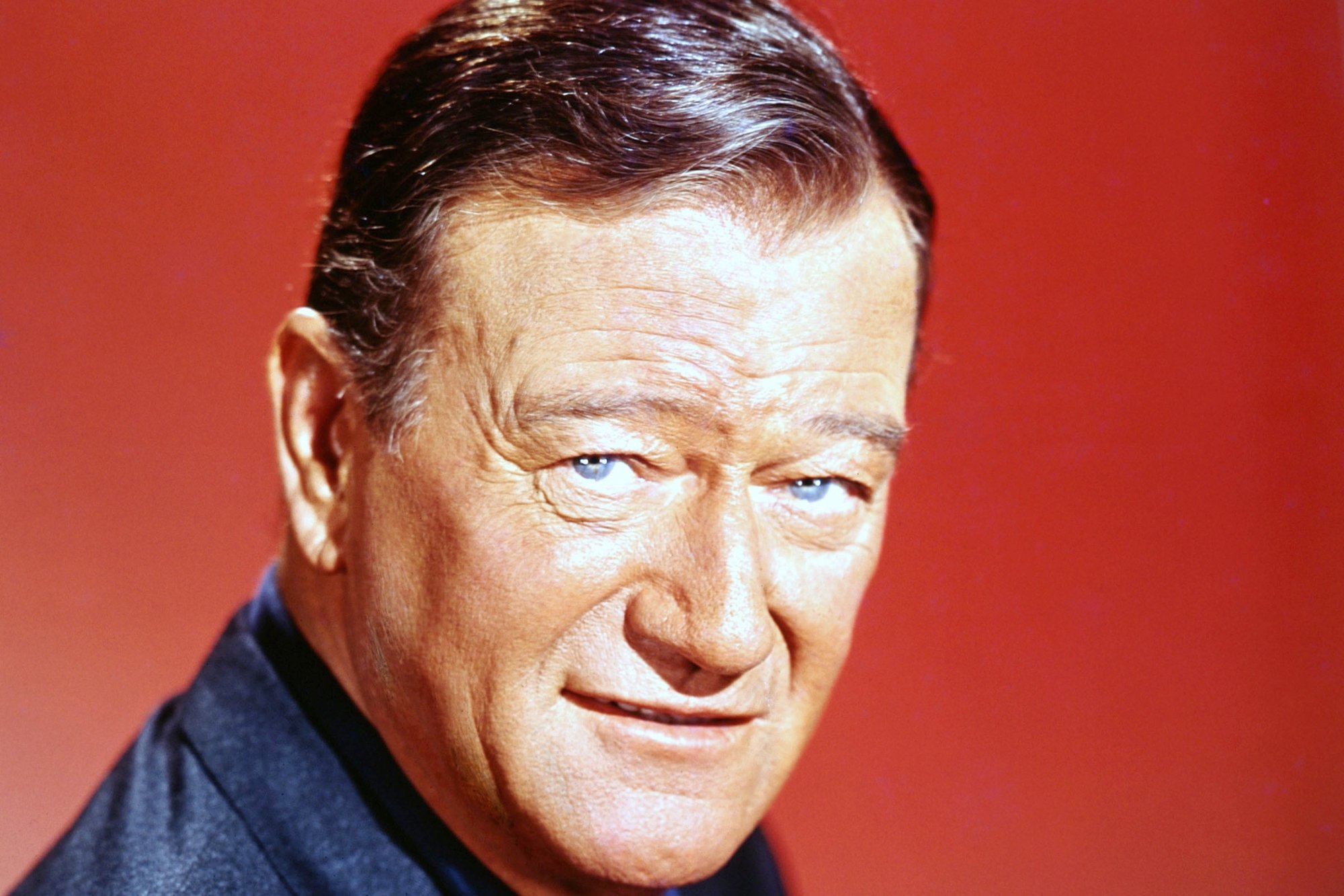 Movie star John Wayne wearing a navy blue jacket and a blue collared shirt underneath in front of a red background