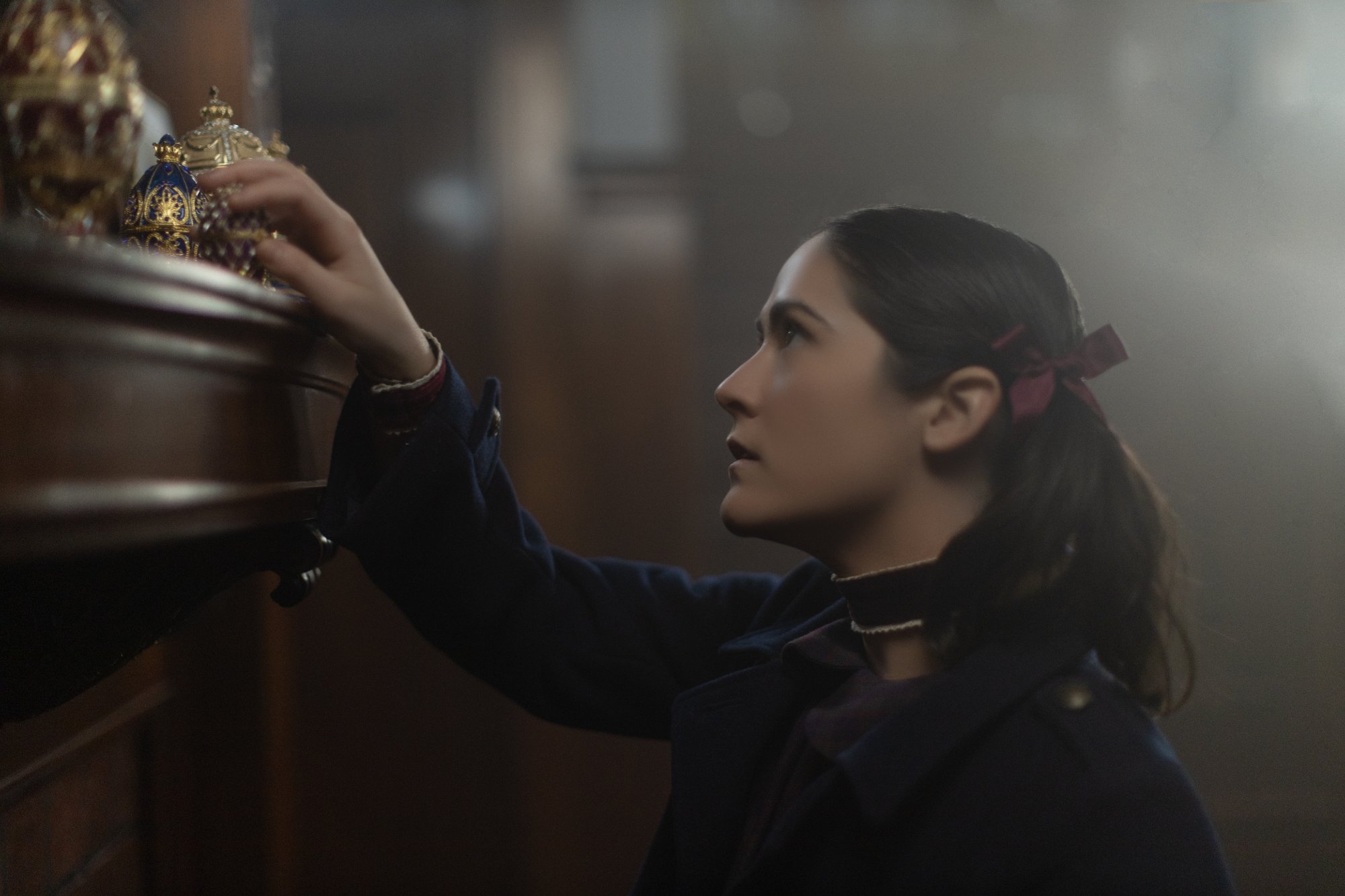'Orphan: First Kill' Isabelle Fuhrman as Esther wearing a blue jacket with red bows in her hair. She's touching a gold ornament on a wooden mantle.