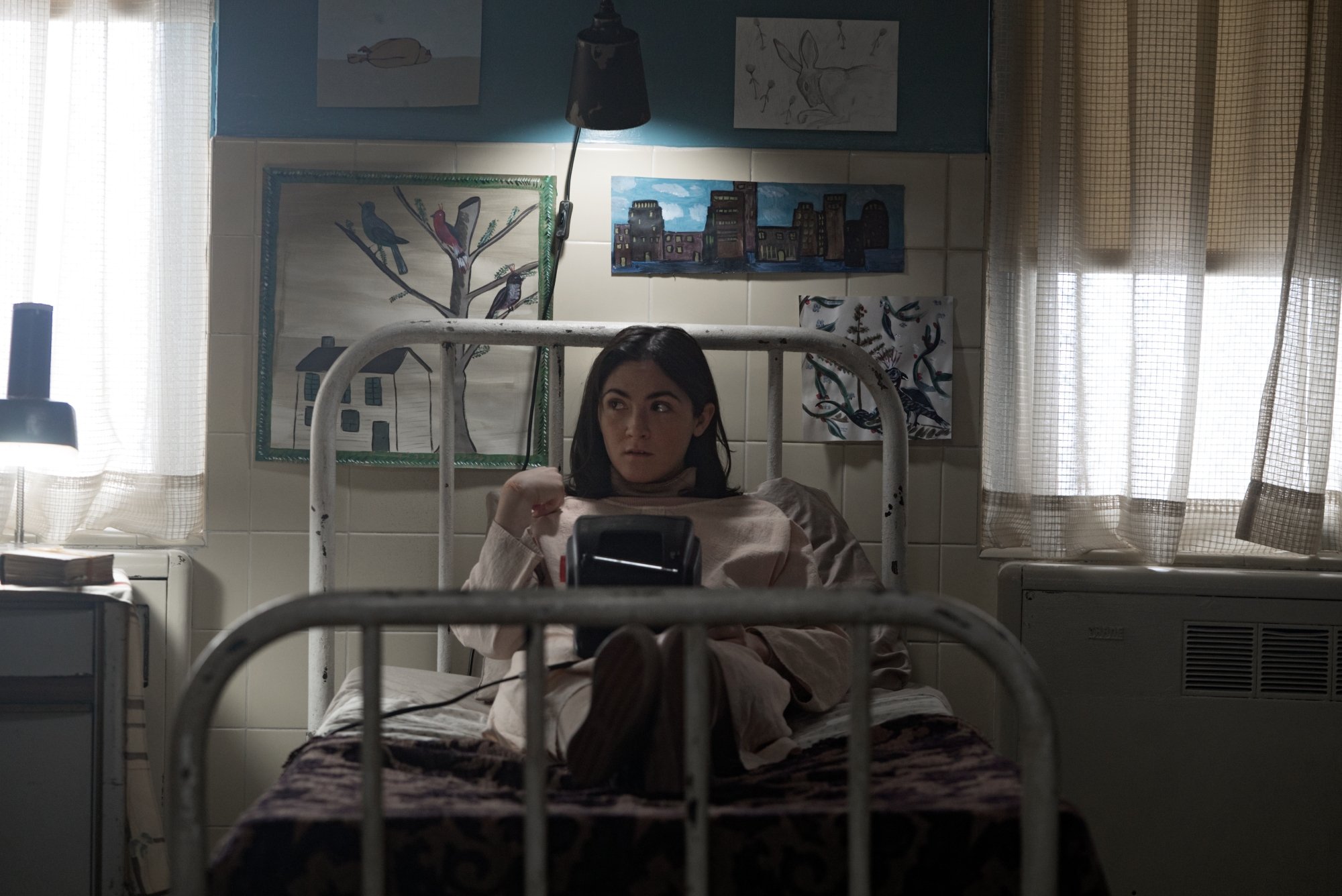 「Orphan: First Kill」のエンディングで、Esther が施設のベッドに座り、後ろの壁に絵が描かれている Isabelle Fuhrman