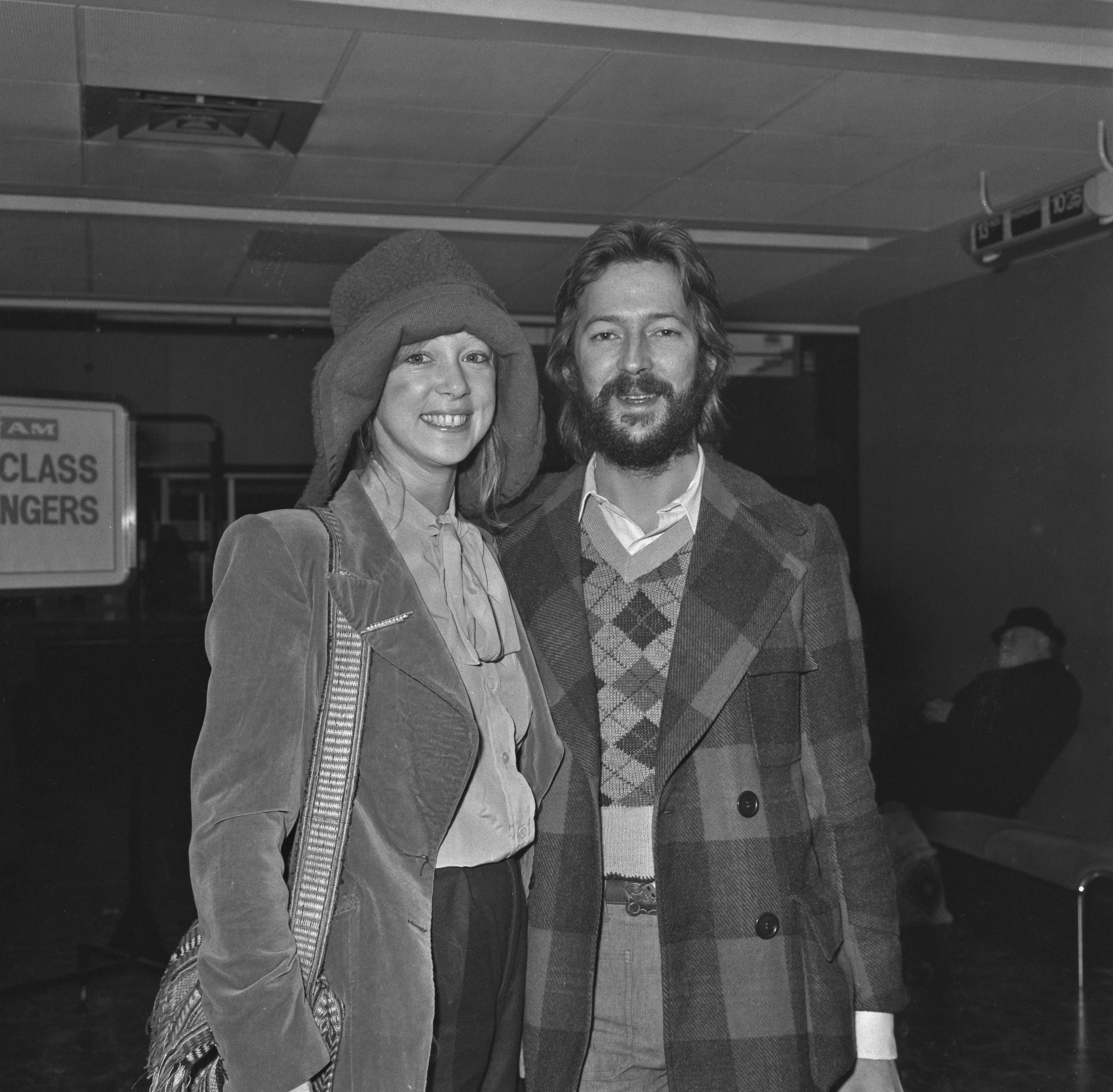 Ein Schwarz-Weiß-Foto von Pattie Boyd, die einen Hut trägt und eine Tasche in der Hand hält und neben Eric Clapton steht. 