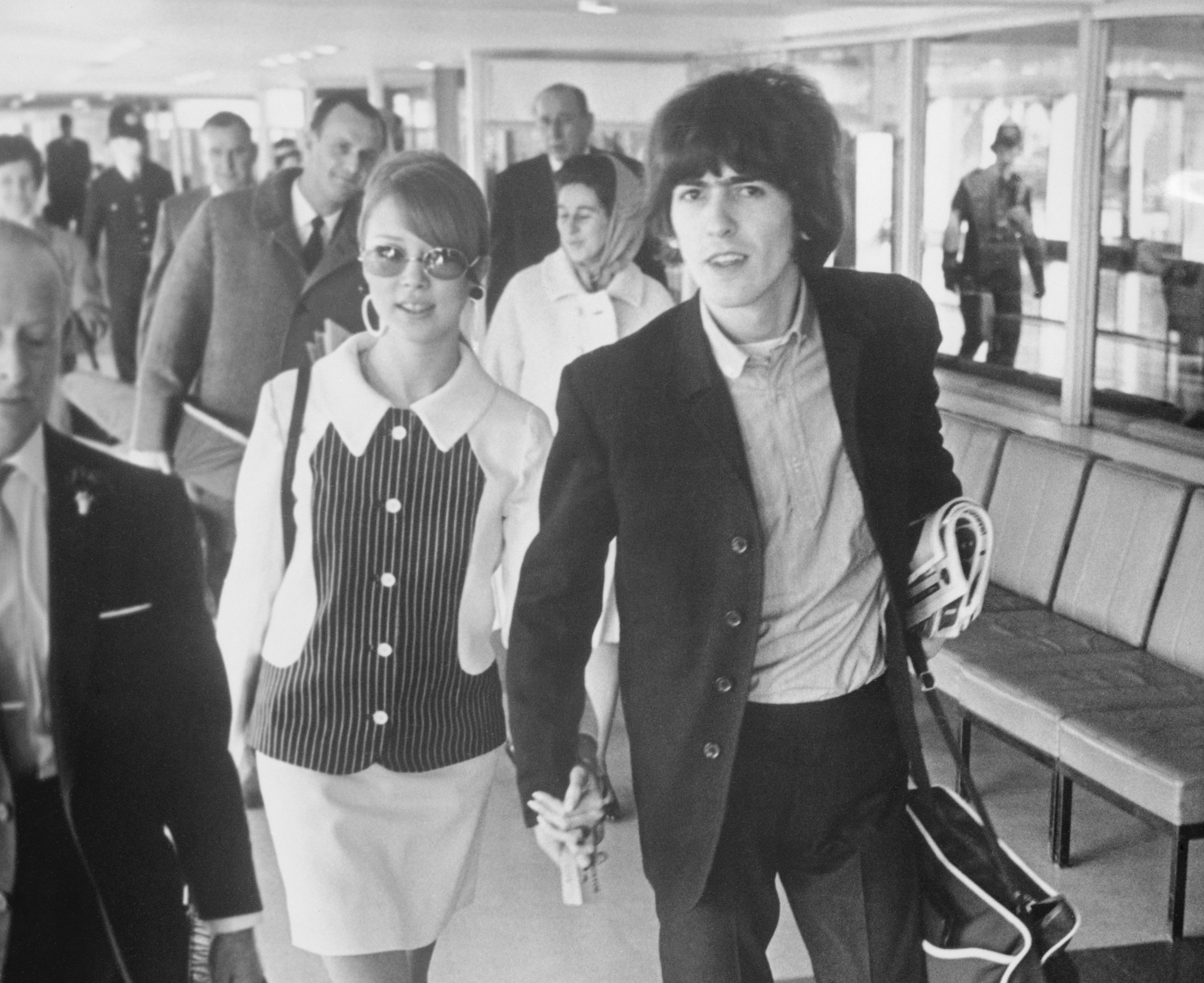 A black and white picture of George Harrison and Pattie Boyd holding hands and walking through an airport.