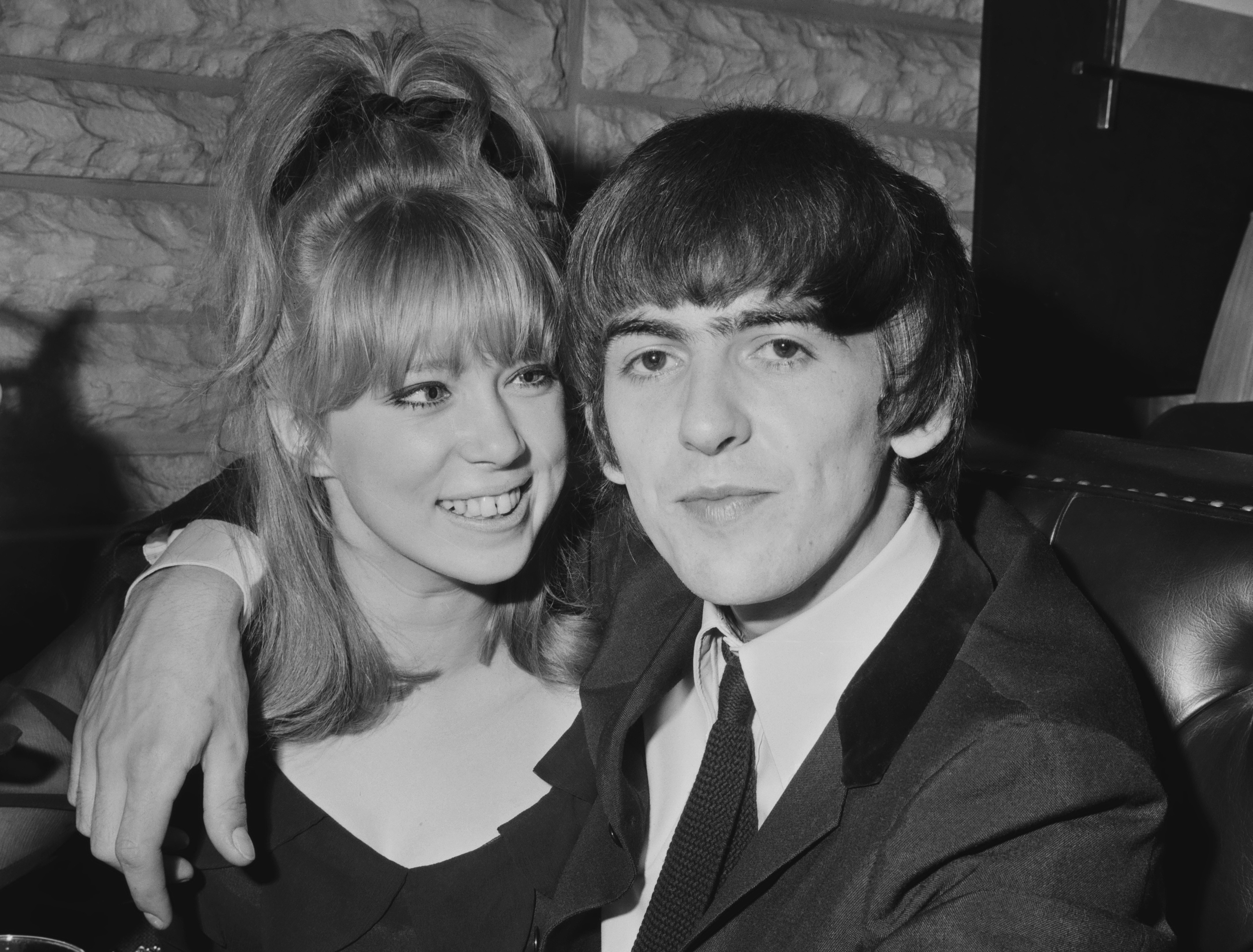 A black and white picture of George Harrison sitting with his arm around Pattie Boyd's shoulders.