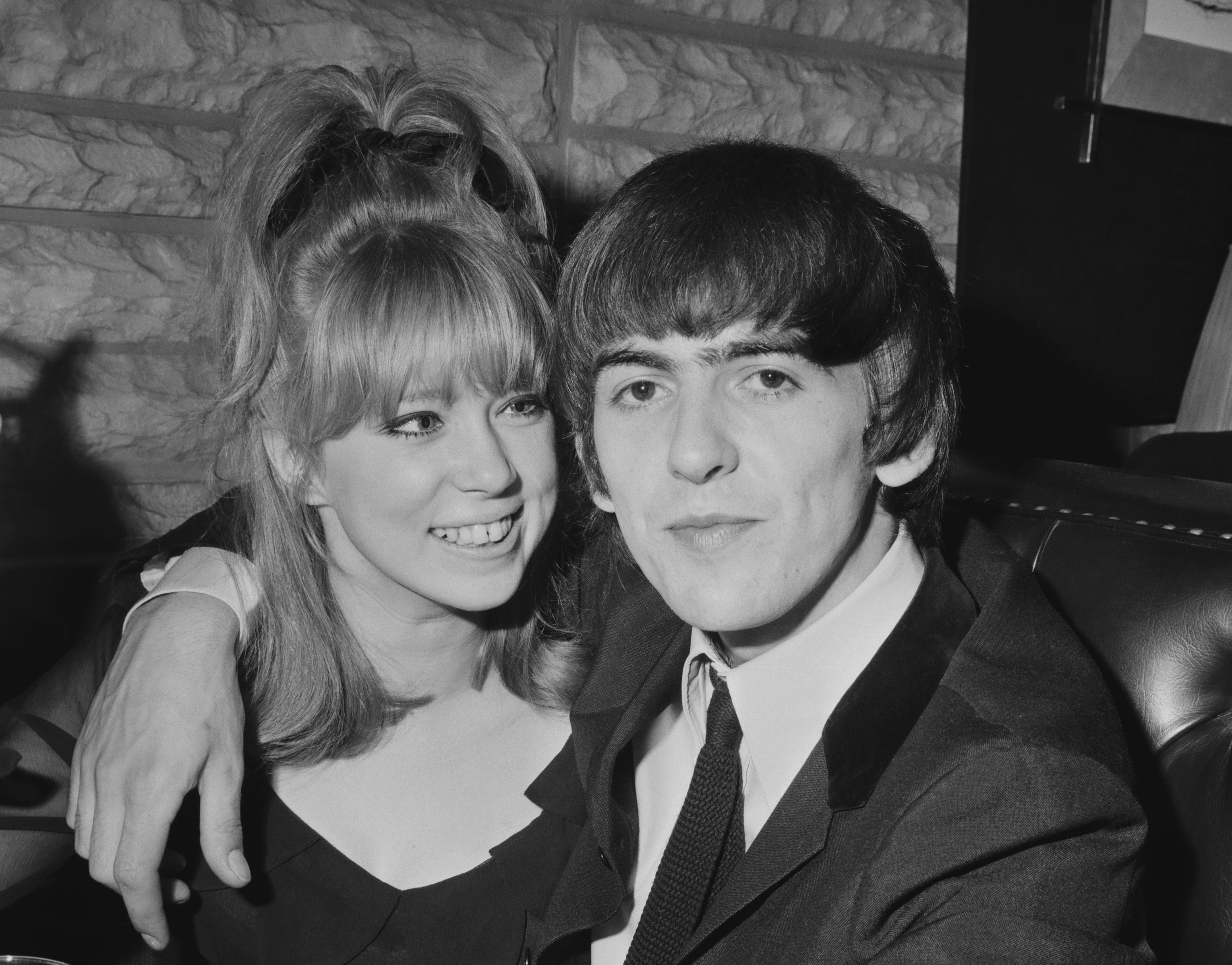 A black and white photo of George Harrison with his arm around Pattie Boyd's shoulder. She stares at him and smiles.
