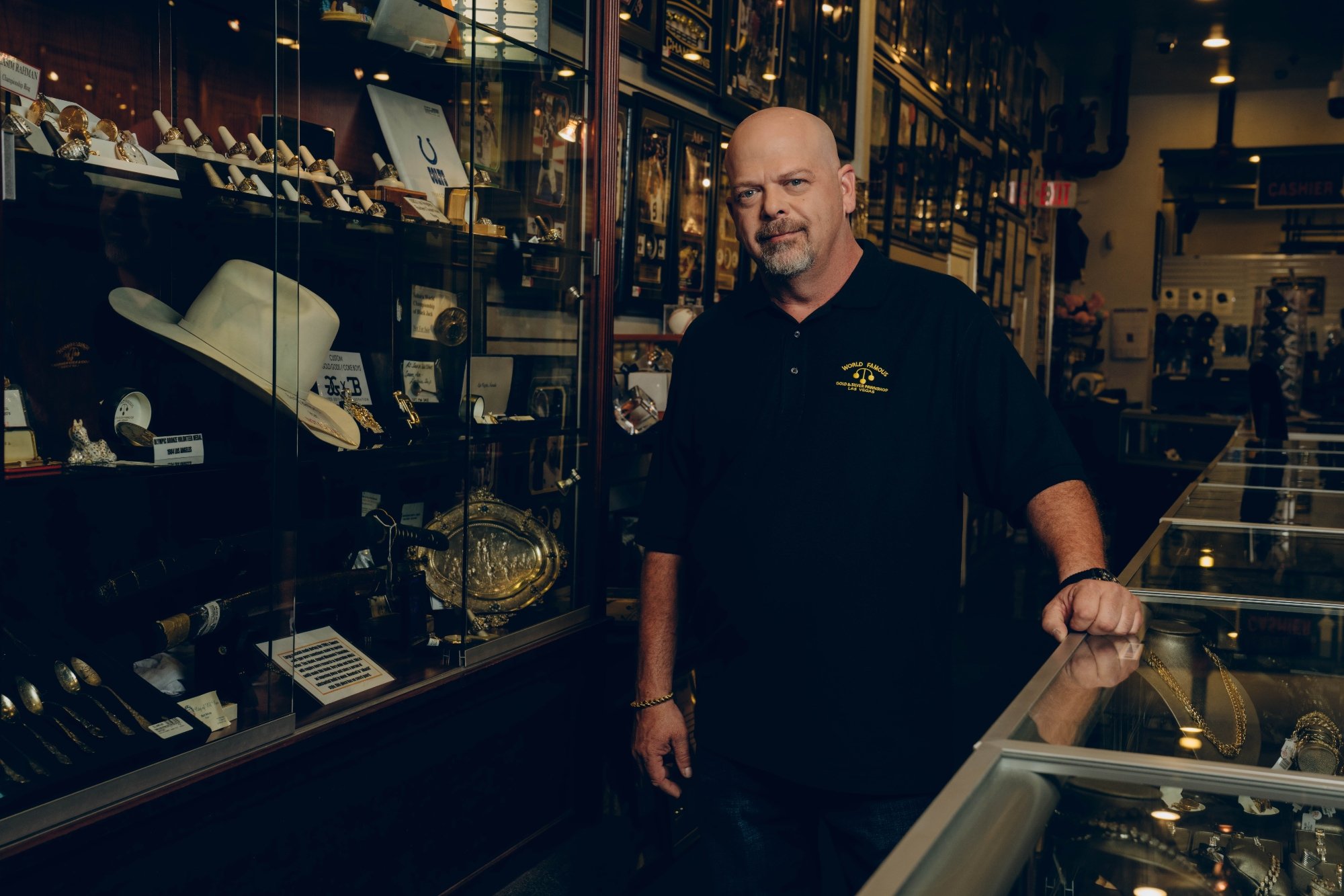 'Pawn Stars' Rick Harrison wearing the shop uniform. He has his hand resting on the glass showcase surrounded by items.