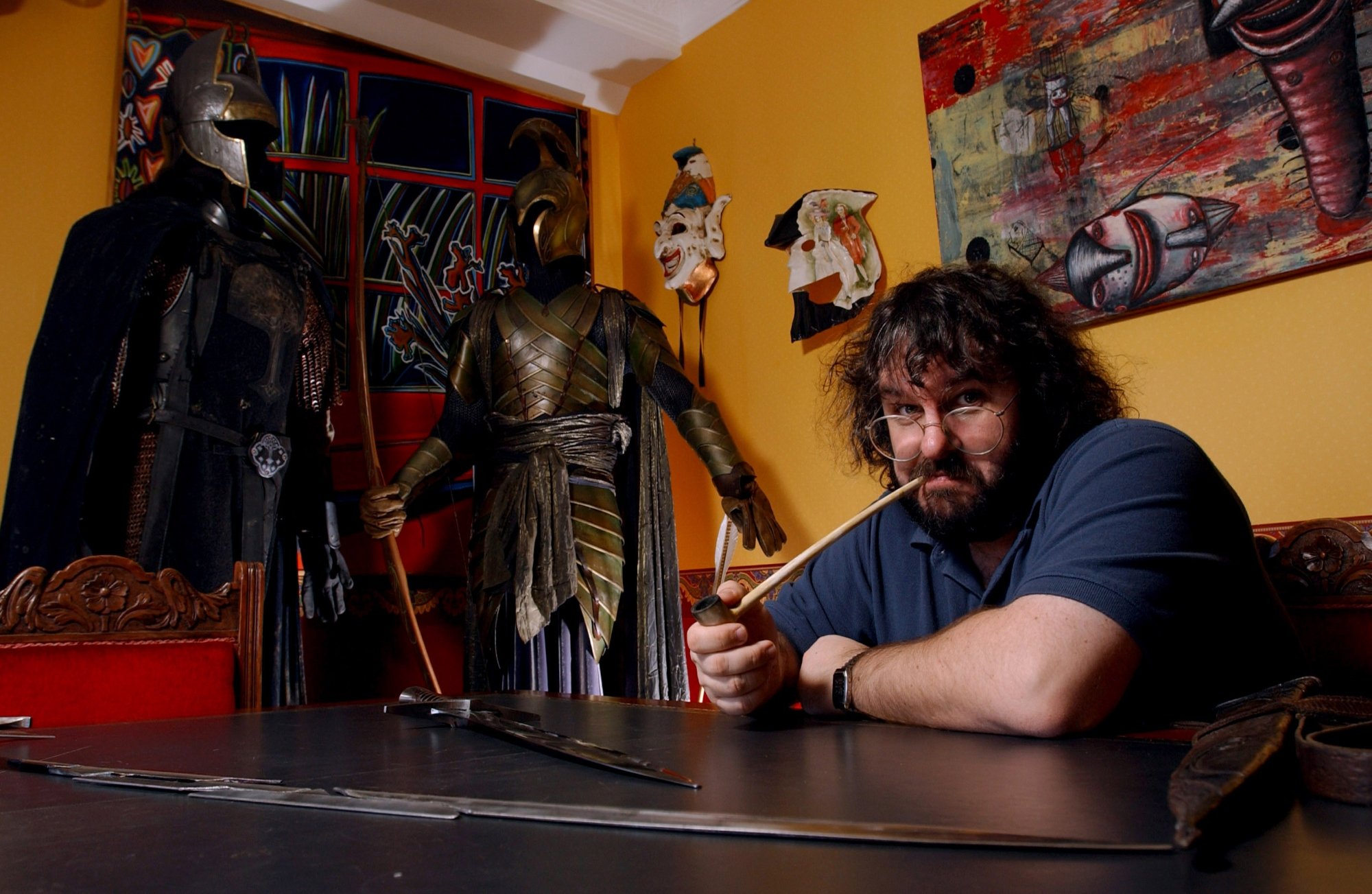Peter Jackson posing with 'The Lord of the Rings' props with a pipe in his mouth. He's wearing a blue shirt and glasses with his arms resting on the table.