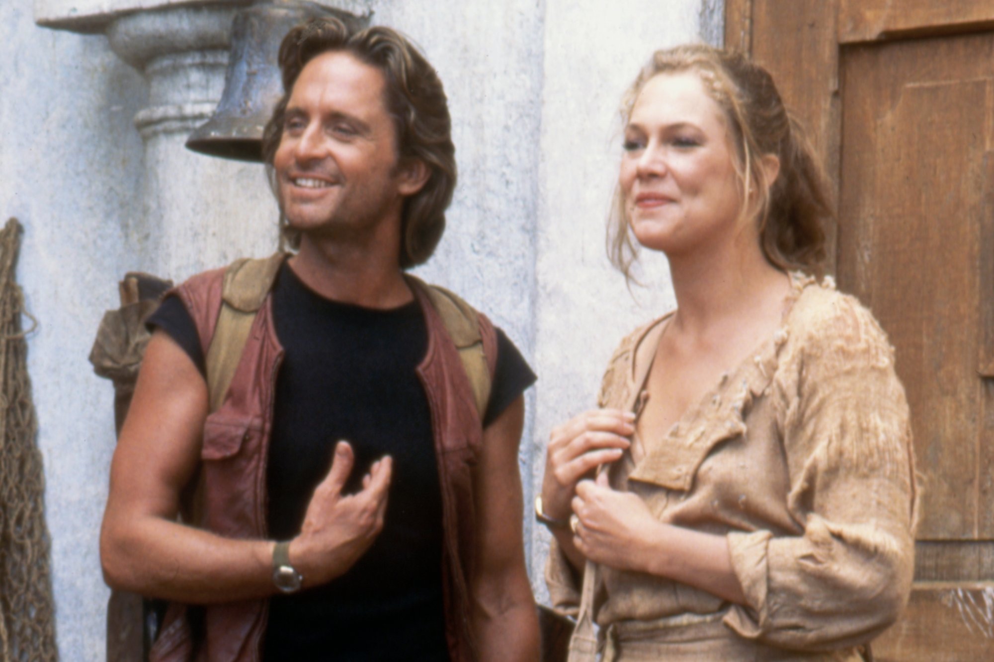 'Romancing the Stone' Michael Douglas as Jack Colton and Kathleen Turner as Joan Wilder. They're smiling in brown adventuring clothing in front of a white wall.