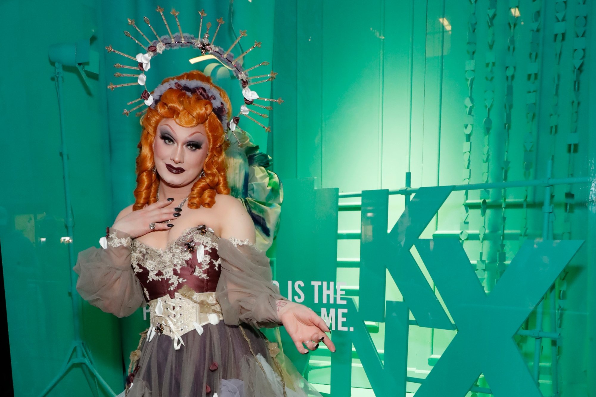 'RuPaul's Drag Race' winner Jinkx Monsoon. They're wearing a fantastical dress with a headpiece in front of a green background.