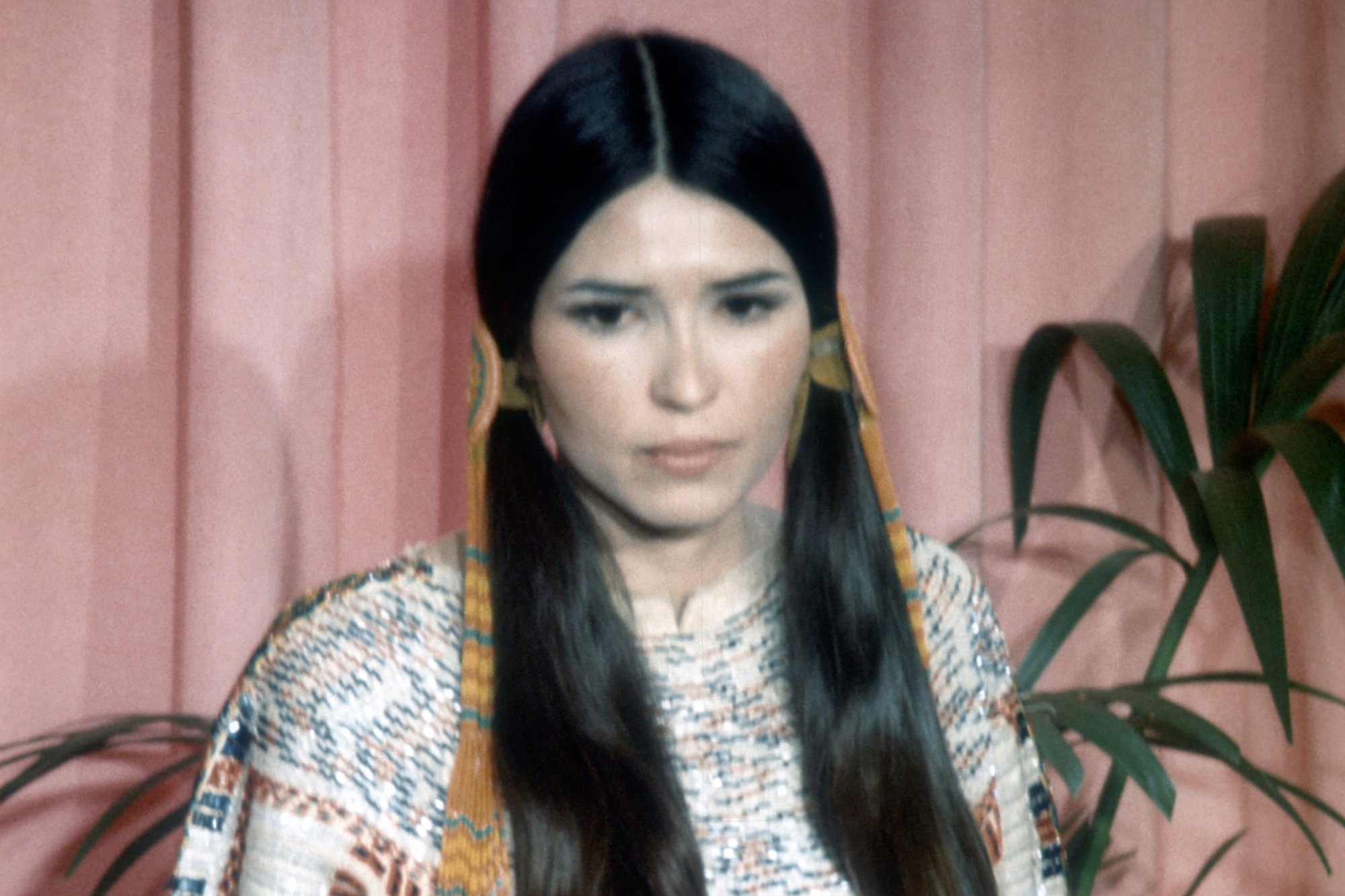 Sacheen Littlefeather at 1973 Oscars wearing traditional Native American dress with a pink curtain and plant behind her