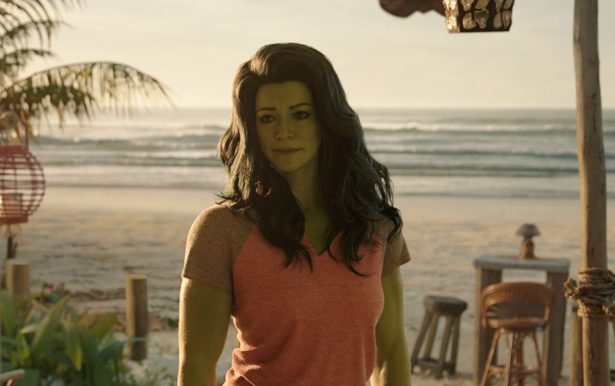 Tatiana Maslany as Jennifer Walters in 'She-Hulk' Episode 1 for our article about its release date and time. She's wearing an orange tank top, has green features, and is standing in front of a beach.