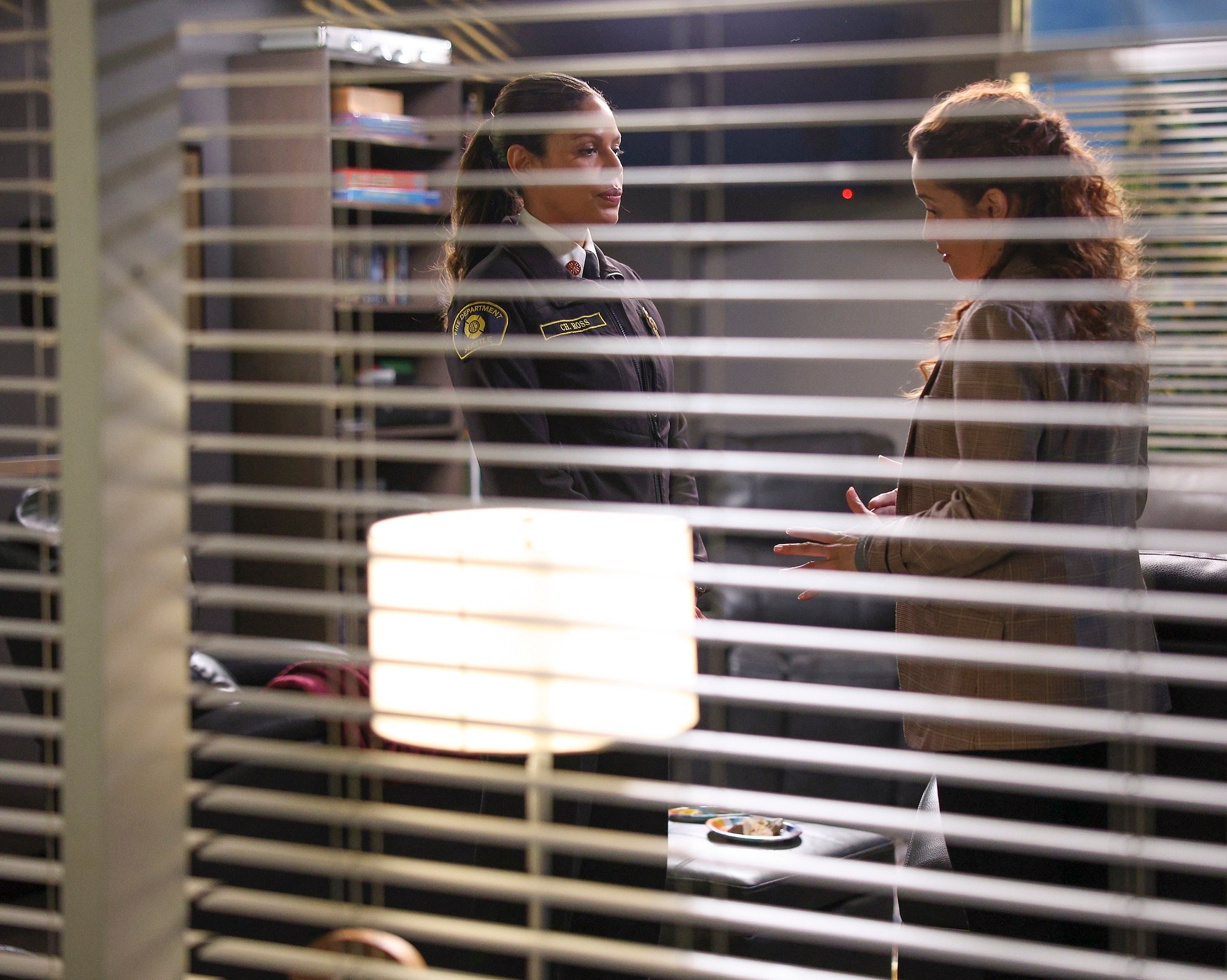 In the 'Station 19' Season 5 finale Merle Dandridge and Jaina Lee Ortiz talk behind open window blinds as Natasha Ross and Andy Herrera