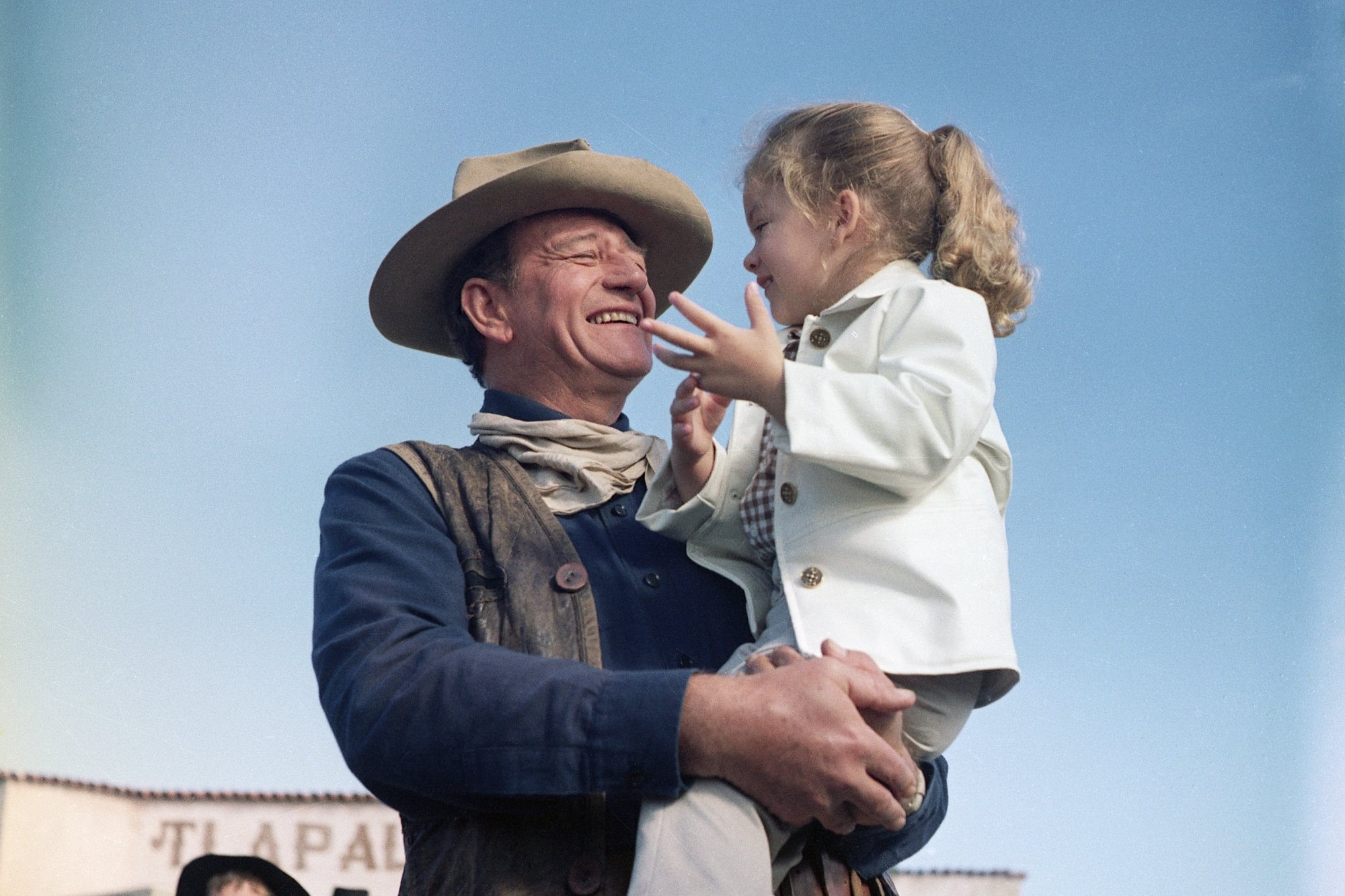 'The Alamo' John Wayne and Aissa Wayne. He holds her in his arms with a big smile on his face. He's wearing a cowboy hat and costume, complete with a scarf and vest.