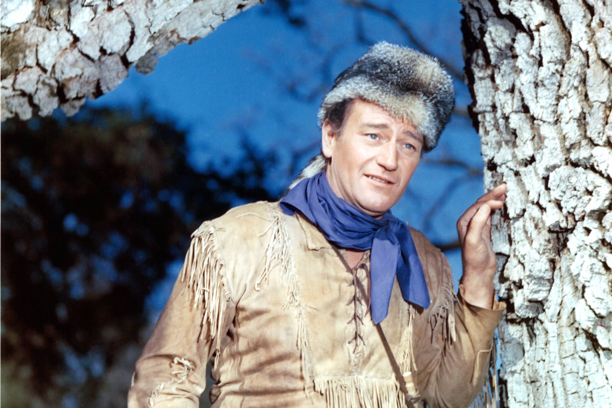 'The Alamo' John Wayne, whose wife was on the set with him. He's wearing an animal pelt hat, fringed Western clothing, while he leans against a tree.