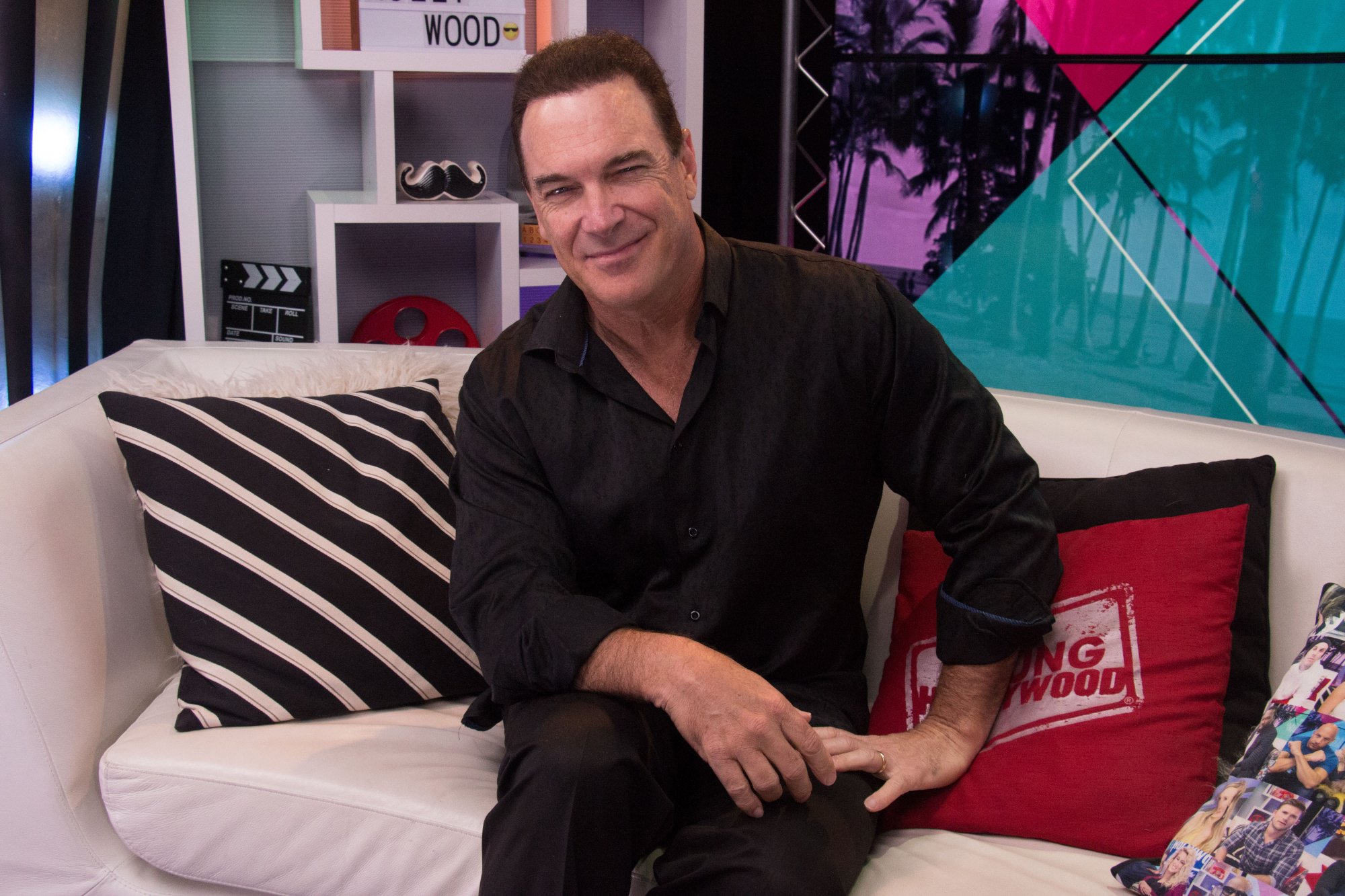 Patrick Warburton, who appeared during 'The Bachelorette' 2022. In the image, he's sitting on a white couch with pink and black pillows. He's wearing all black and smiling.