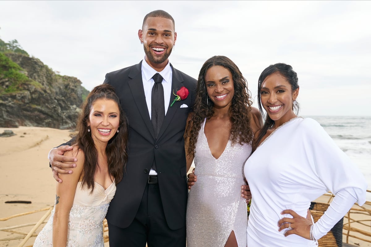 Kaitlyn Bristowe, Nayte Olukoya, Michelle Young, and Tayshia Adams in The Bachelorette Season 18, pose for a photo on the beach. The women wear white and Nayte wears a suit.