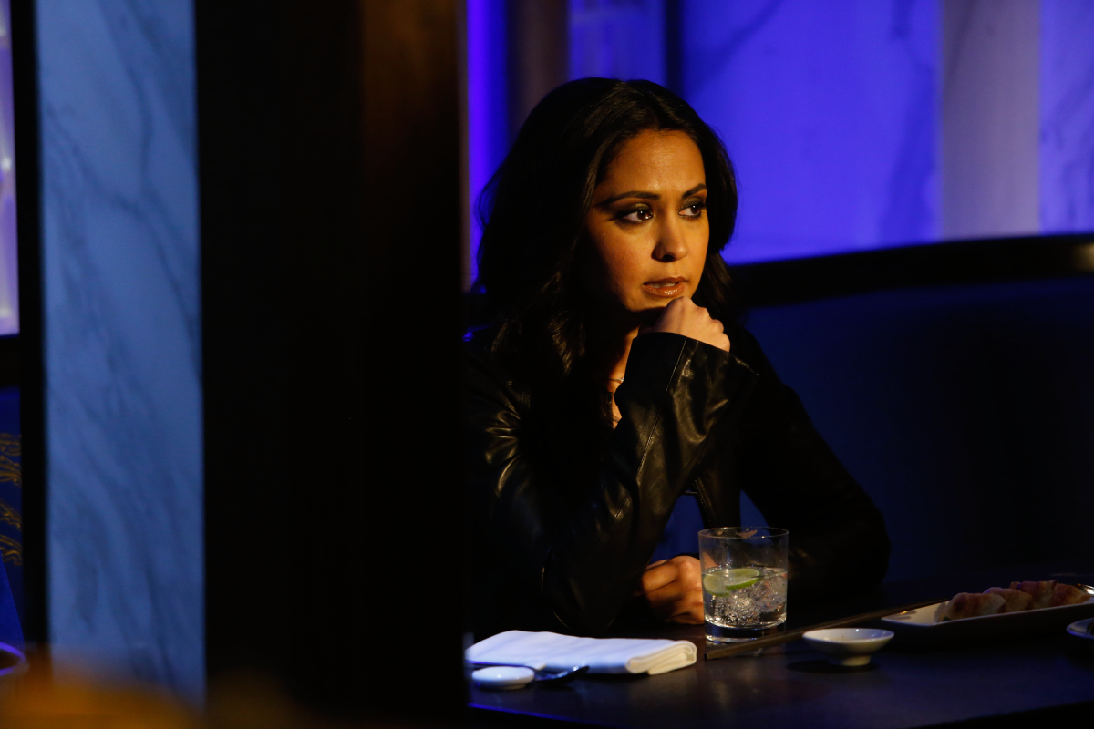 Meera Malik's daughter will appear in The Blacklist Season 10. Meera sits at a table with a cocktail in front of her.