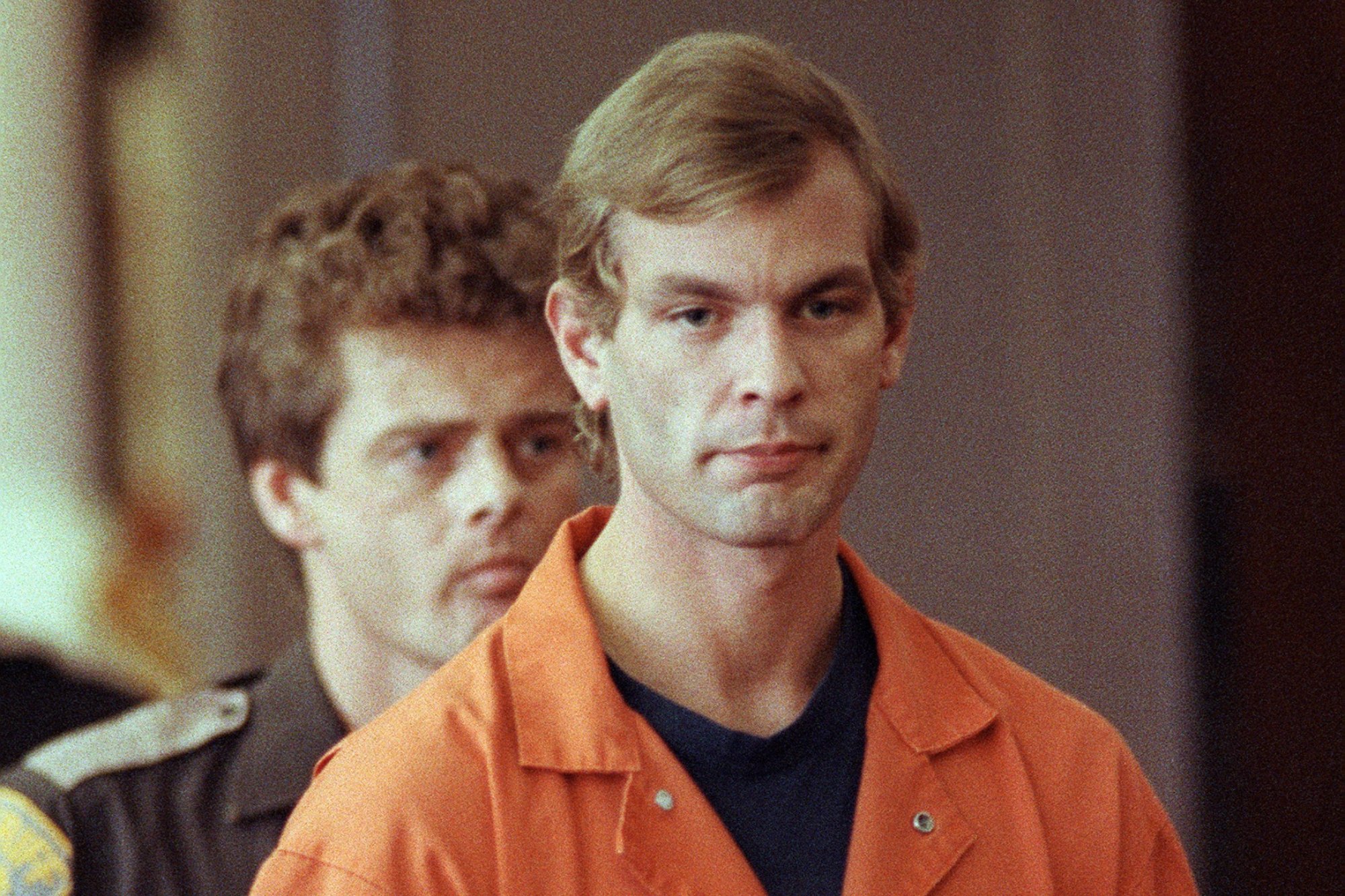 'The Jeffrey Dahmer Tapes' release date revealed.  He is wearing an orange prison jumpsuit with a serious look on his face and a security guard standing behind him.
