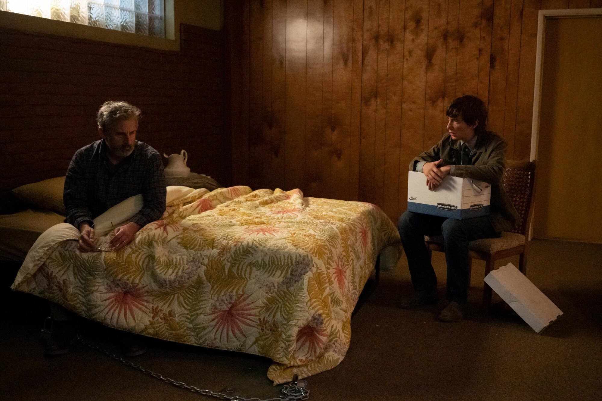 Steve Carell and Domhnall Gleeson in 'The Patient,' which will drop episode 3 on Sept. 6. Carell's character is sitting beneath the covers in bed, and Gleeson's character is sitting on a chair next to the bed and holding a box.