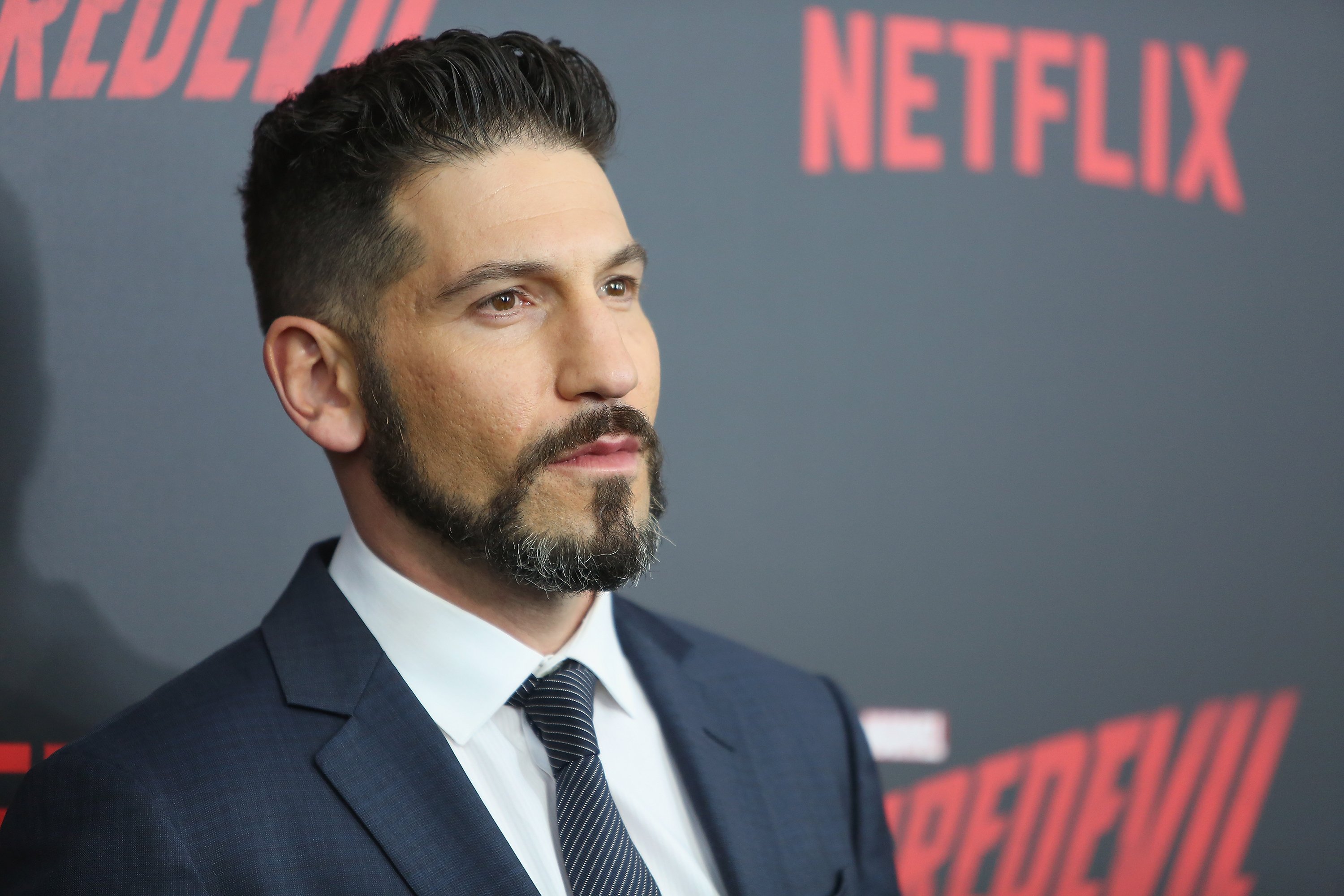 Jon Bernthal, who played the Punisher in the MCU, wears a black suit over a white button-up shirt and black and white striped tie.