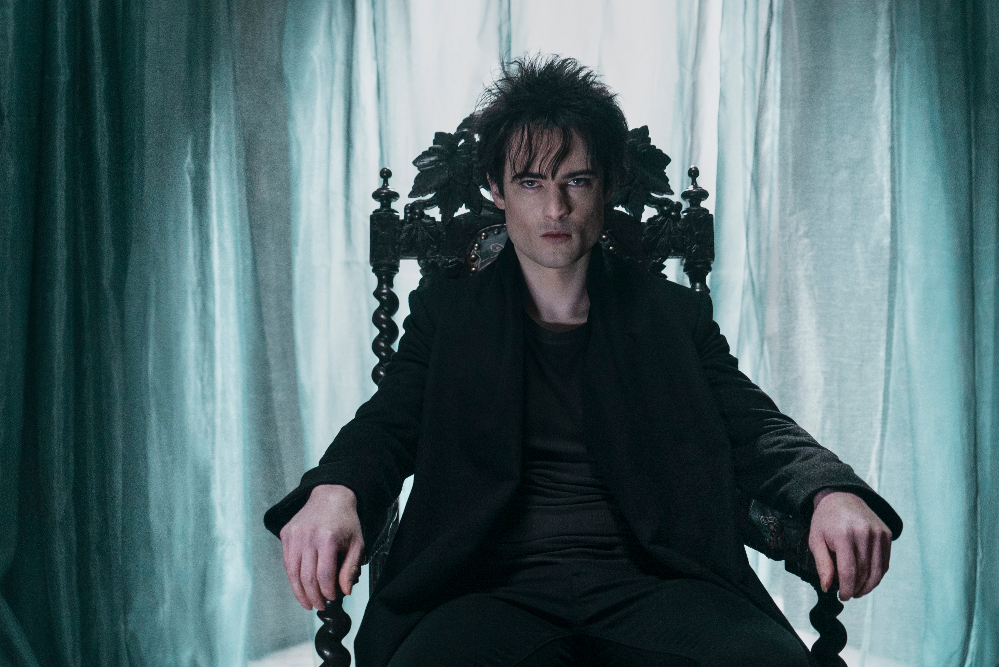Tom Sturridge as Dream in the cast of Netflix's 'The Sandman.' He's sitting in a chair with light blue curtains behind him.