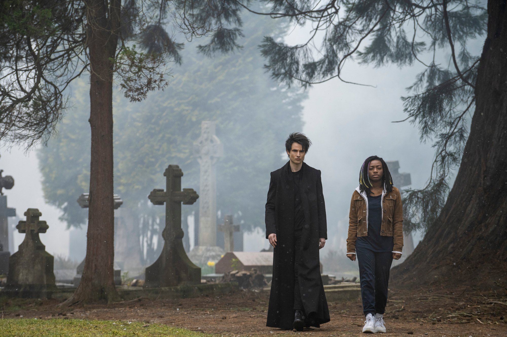 Tom Sturridge and Kyo Ra as Morpheus and Rose Walker in 'The Sandman' for our review of the Netflix series. The characters are walking side by side in a graveyard.