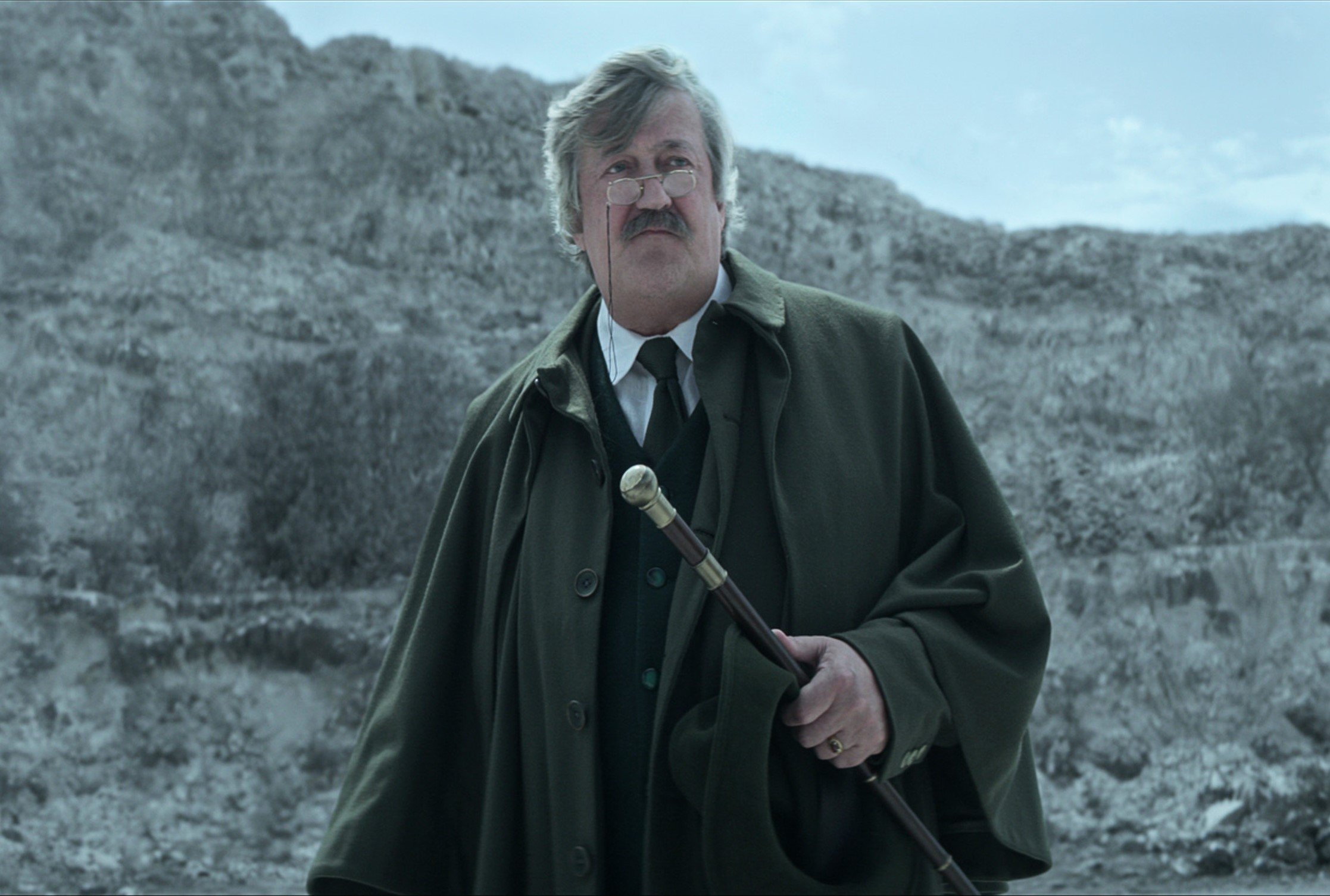 Stephen Fry as Gilbert in the cast of 'The Sandman.' He's holding a cane and standing before mountains.