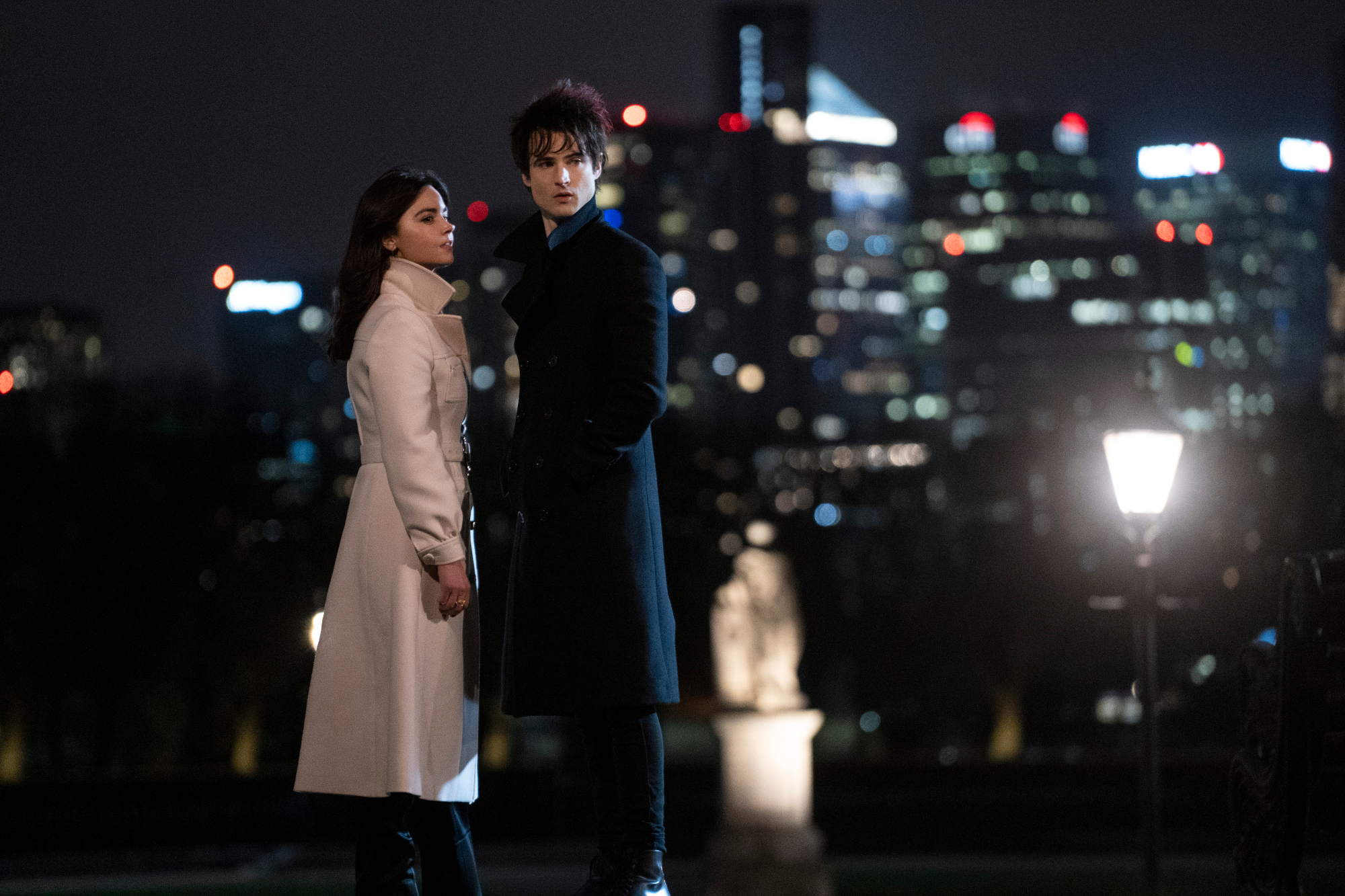 Jenna Coleman as Johanna and Tom Sturridge as Dream in 'The Sandman,' which will drop its first 10 episodes on Netflix on Aug. 5. The pair are standing next to one another and looking at something off-screen. The city is behind them.
