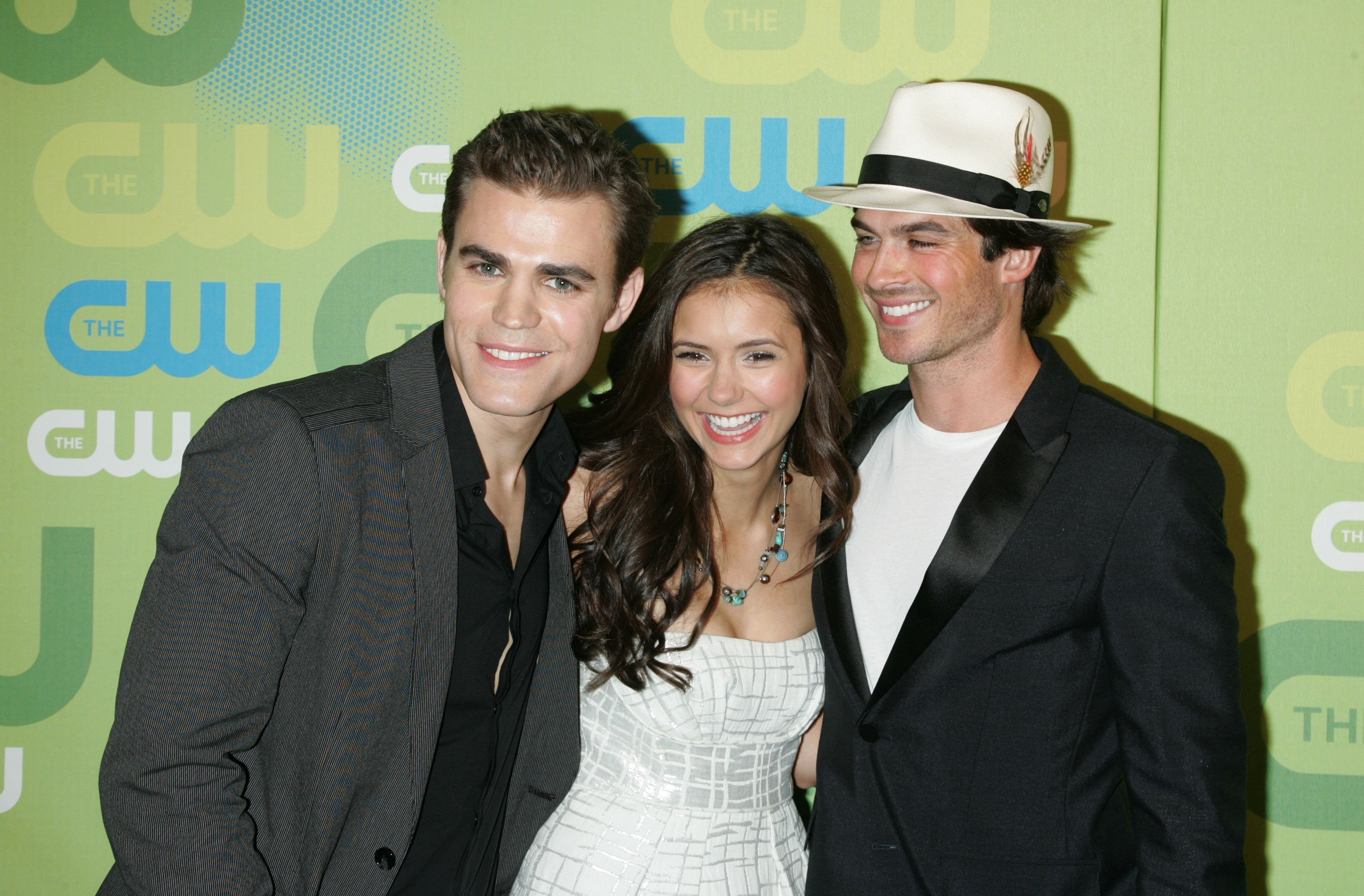 Paul Wesley, Nina Dobrev, and Ian Somerhalder, who starred on 'The Vampire Diaries,' which is leaving Netflix in September 2022, pose for pictures together on the red carpet. Wesley wears a gray suit over a black button-up shirt. Dobrev wears a white strapless dress. Somerhalder wears a black suit over a white shirt and a white fedora.