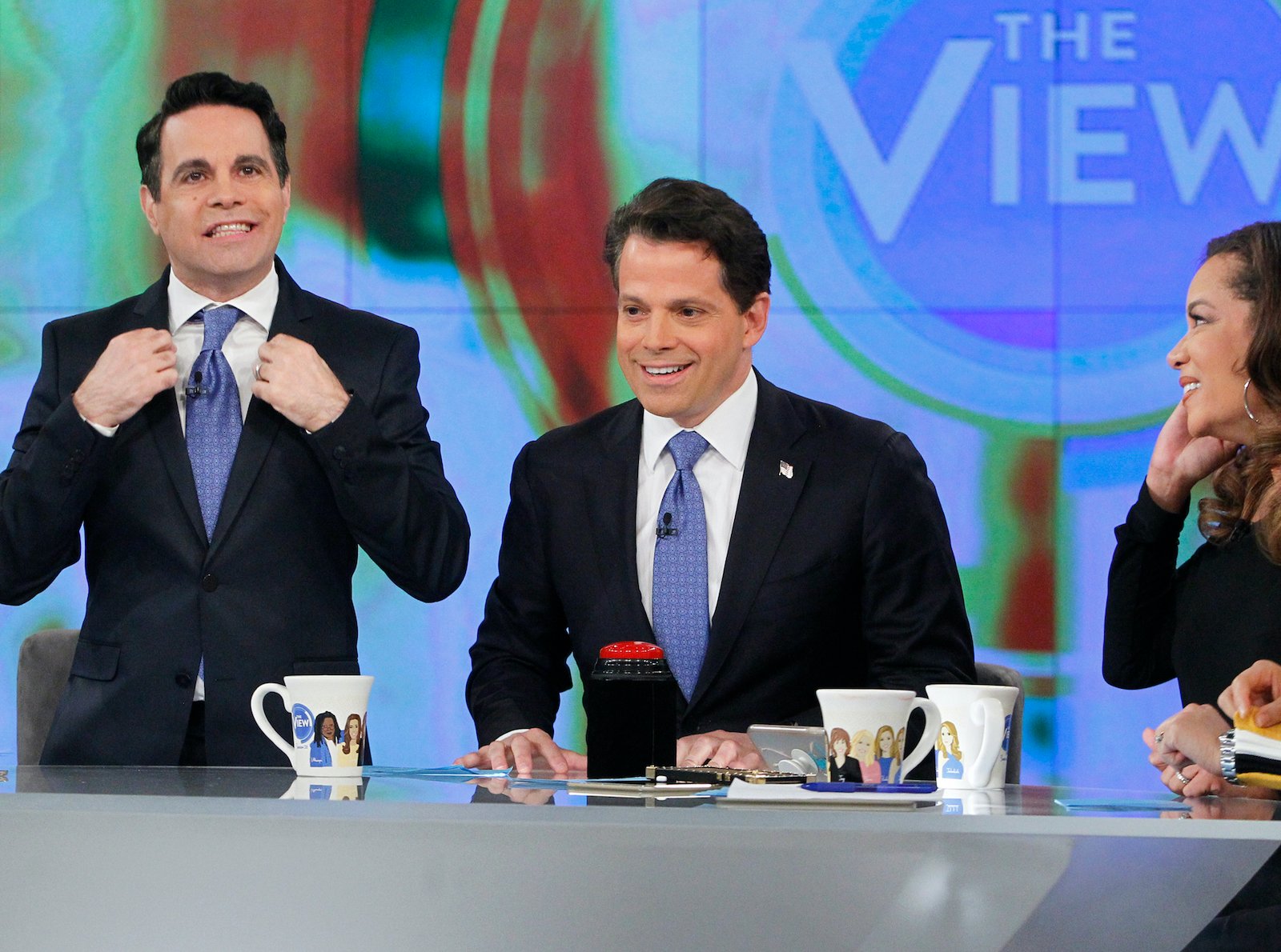 Mario Cantone holds his jacket lapel while standing next to Anthony Scaramucci and Sunny Hostin who are seated
