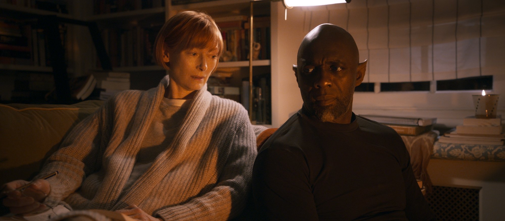 'Three Thousand Years of Longing' Tilda Swinton as Alithea and Idris Elba as The Djinn sitting next to each other on the couch side-eyeing each other with a bookcase behind them