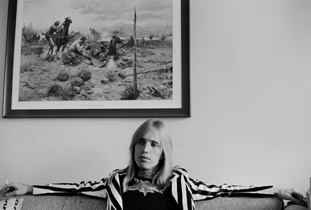 A black and white picture of Tom Petty wearing a striped jacket and sitting on a couch underneath a painting. 