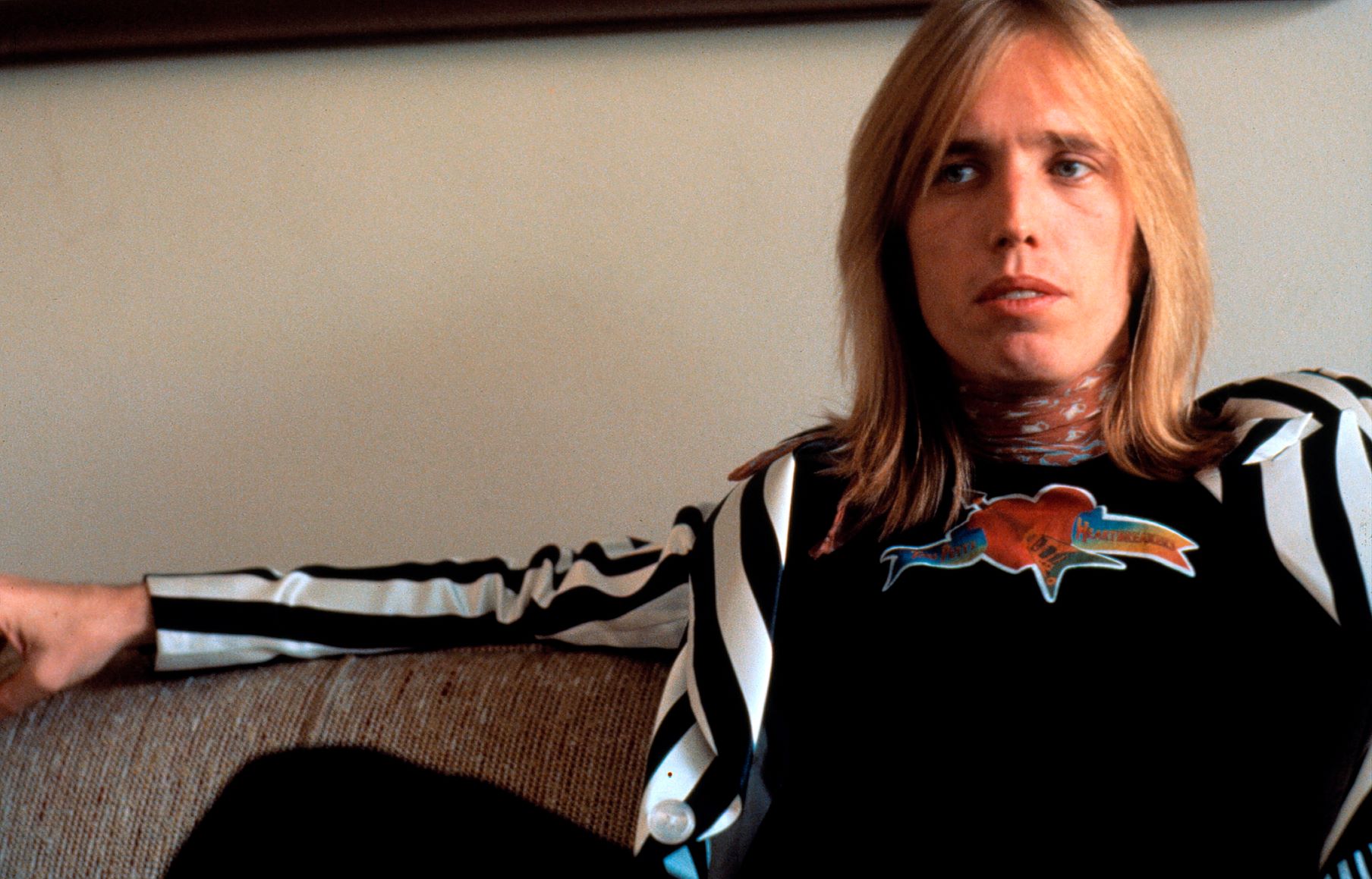 Tom Petty wears a black and white jacket and sits on a couch.