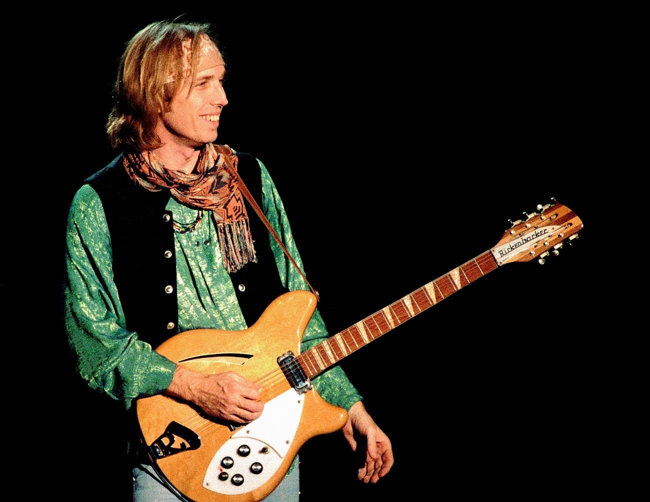 Tom Petty wears a green shirt and vest and holds a guitar. 