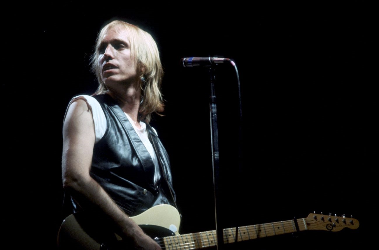 Tom Petty wears a leather vest and holds a guitar. He stands in front of a microphone. 