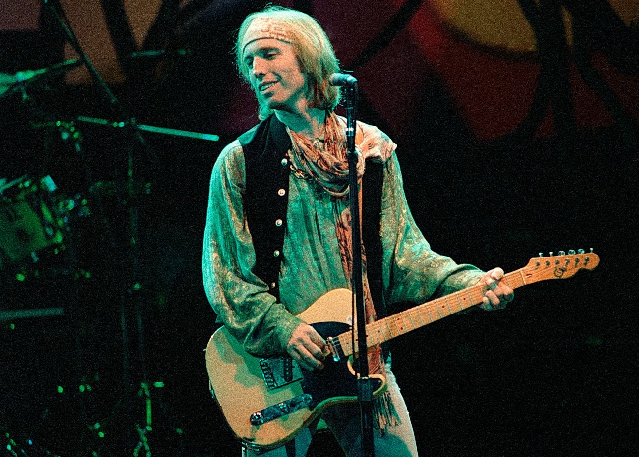 Tom Petty wears a green shirt and plays guitar. He stands in front of a microphone.