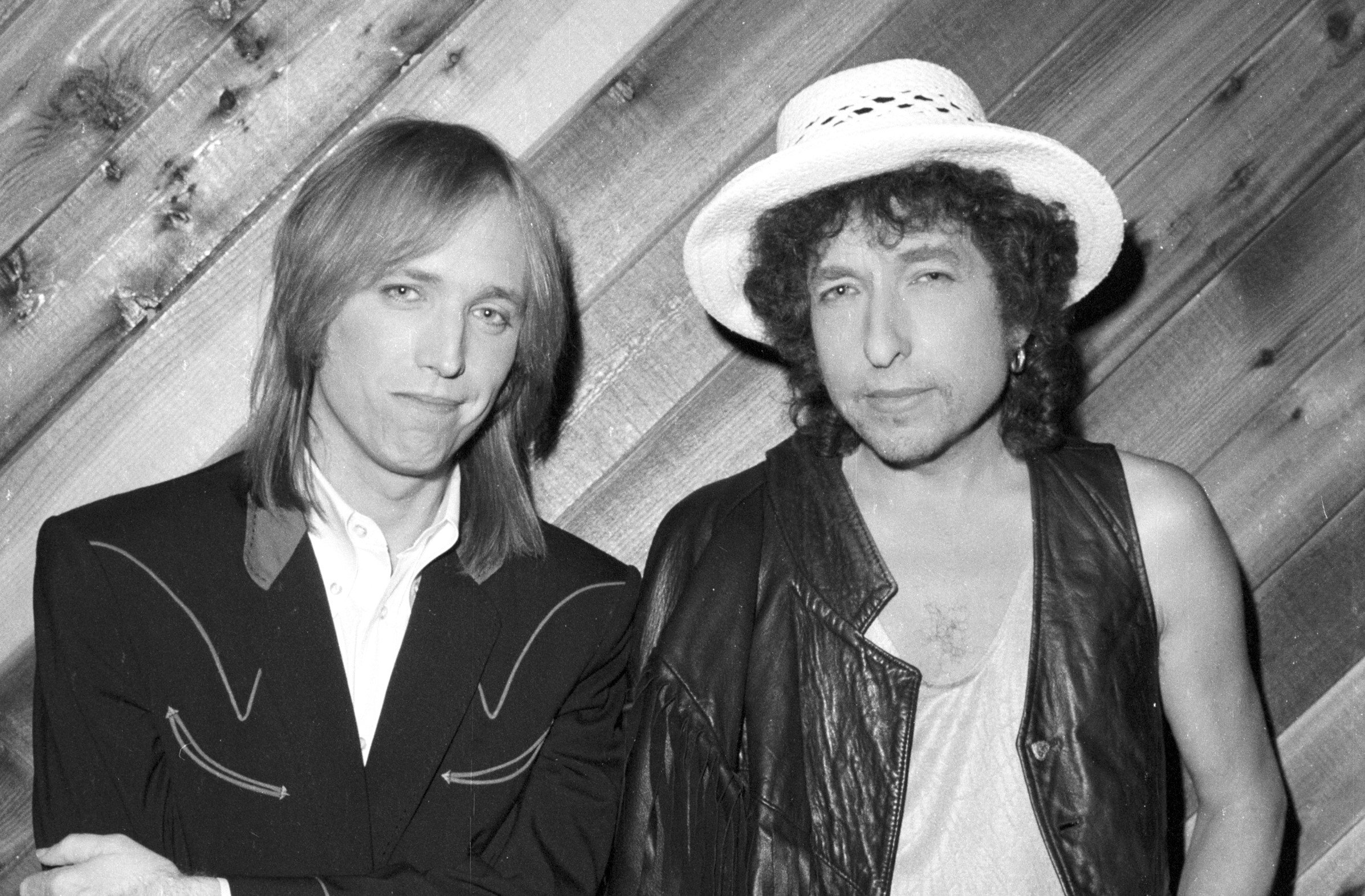 A black and white picture of Tom Petty and Bob Dylan standing against a wood paneled wall.