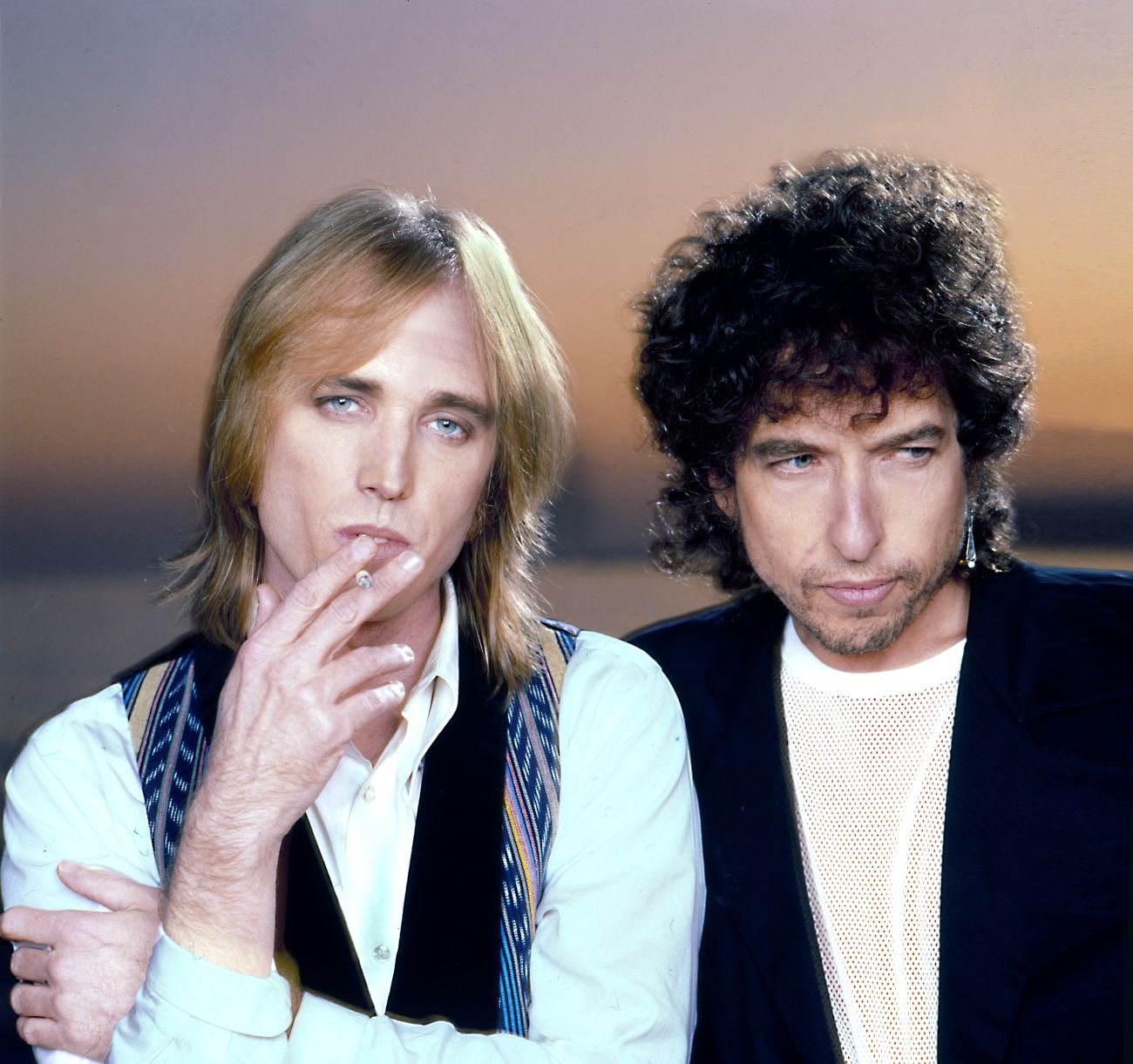 Tom Petty and Bob Dylan stand in front of a sunset background. Petty holds a cigarette.