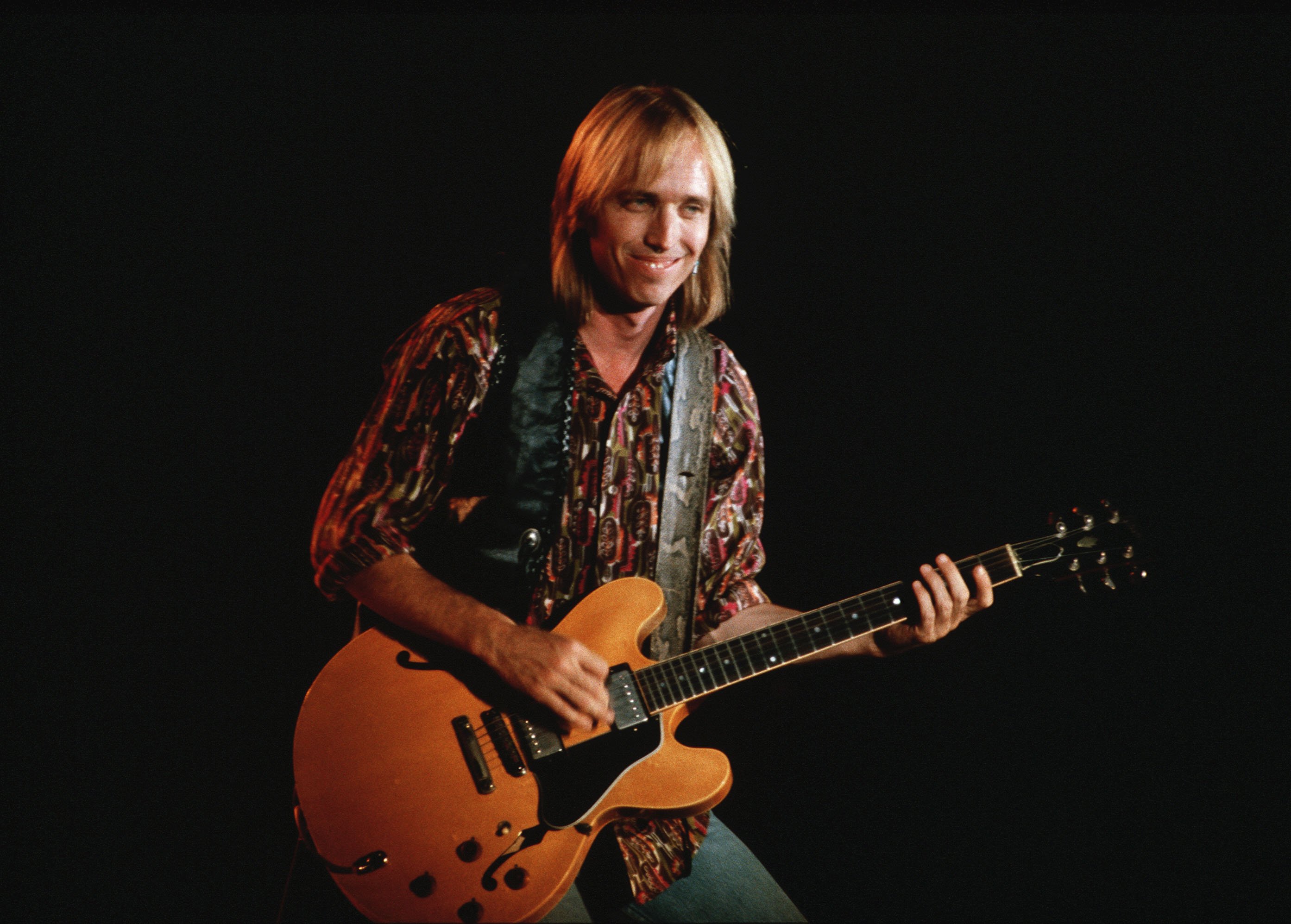 Tom Petty wears a vest and holds a guitar. 