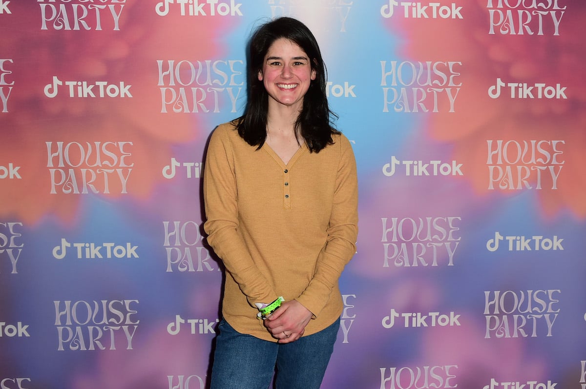 TikTok star Elyse Myers in a yellow shirt and jeans posing in front of a TikTok House Party backdrop